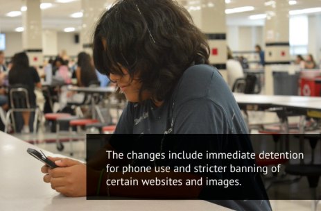 A student looks down at a cell phone at a cafeteria table.