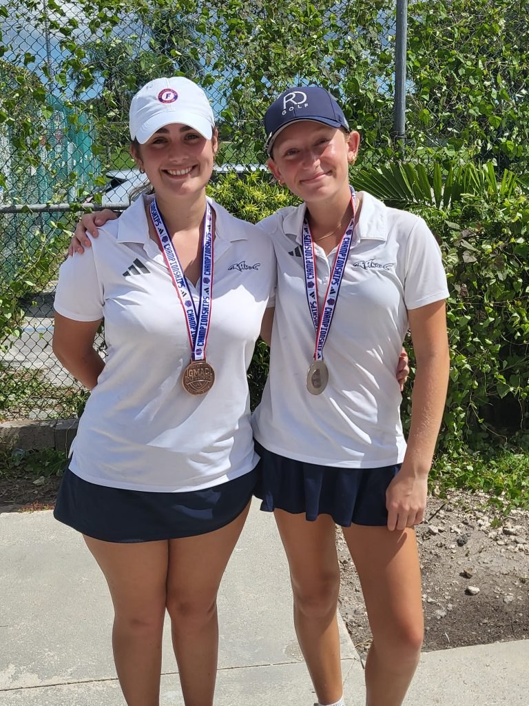 Two young women in white golf uniforms each wearing a competition medal pose outdoors.