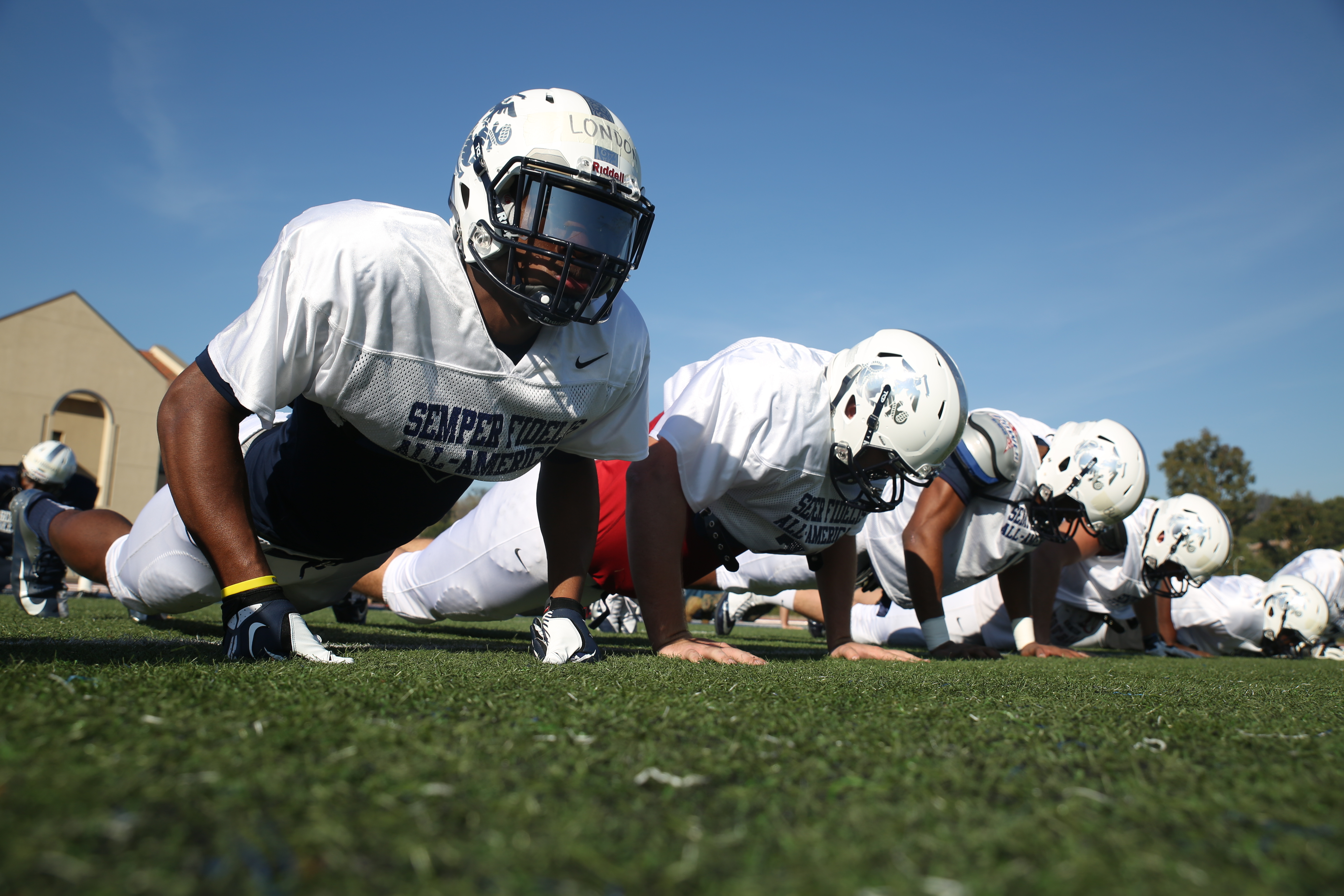 Football players in white helmets and pads doing push-ups in a line on a grass field.