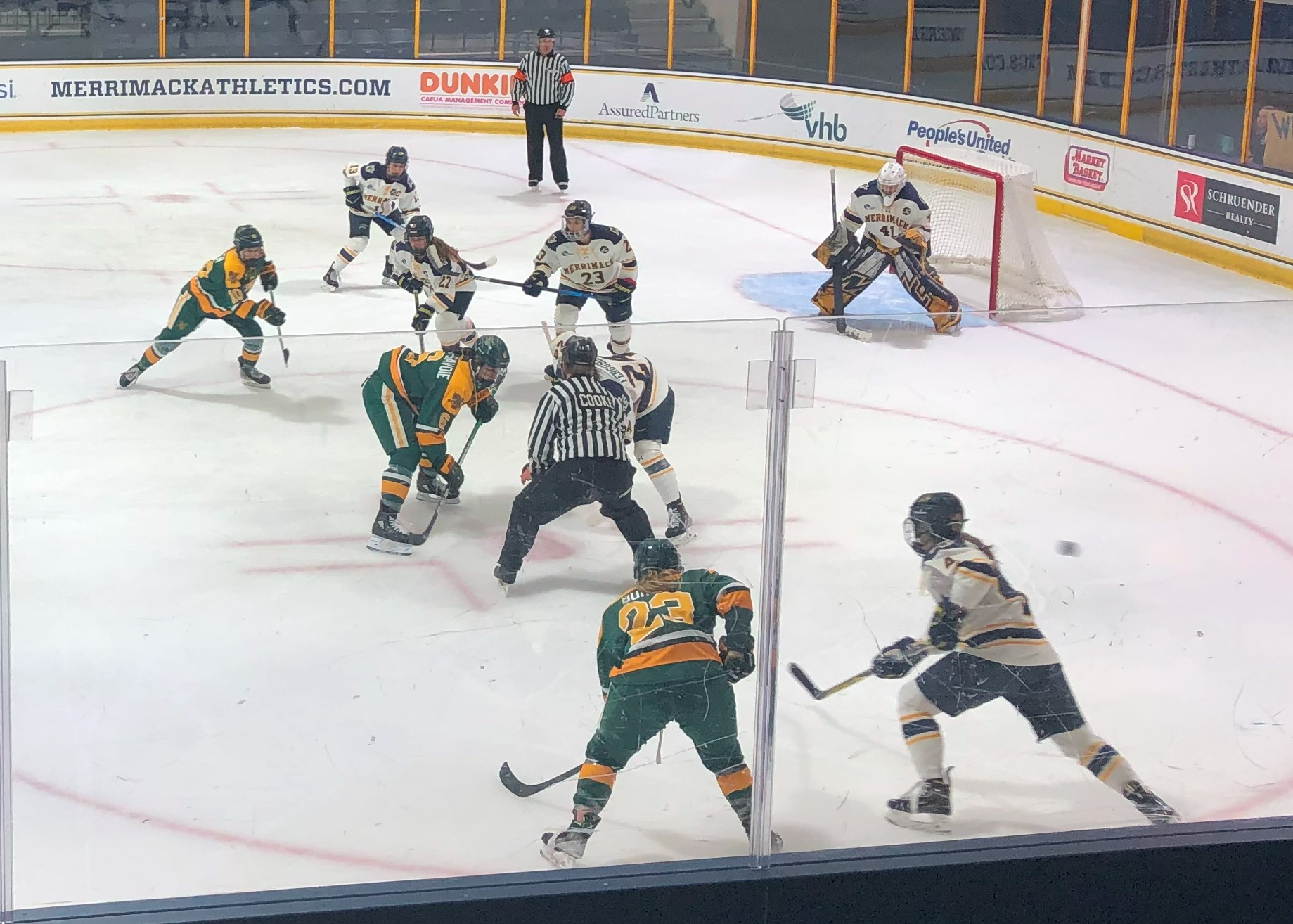 Ice hockey players in green and gold uniforms compete near the goal net at an indoor rink.