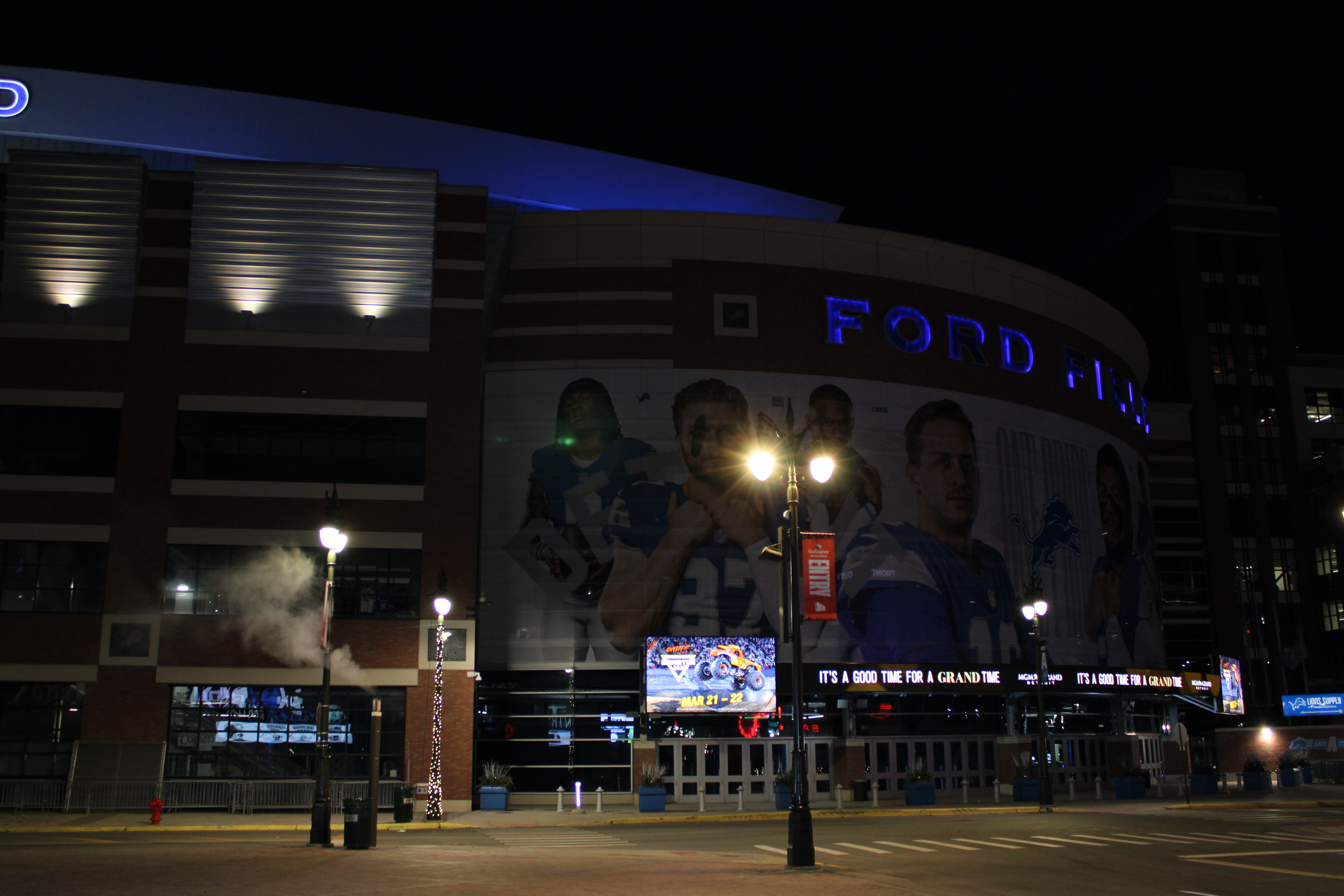 Ford Field exterior at night with illuminated blue signage and large player murals on the facade.