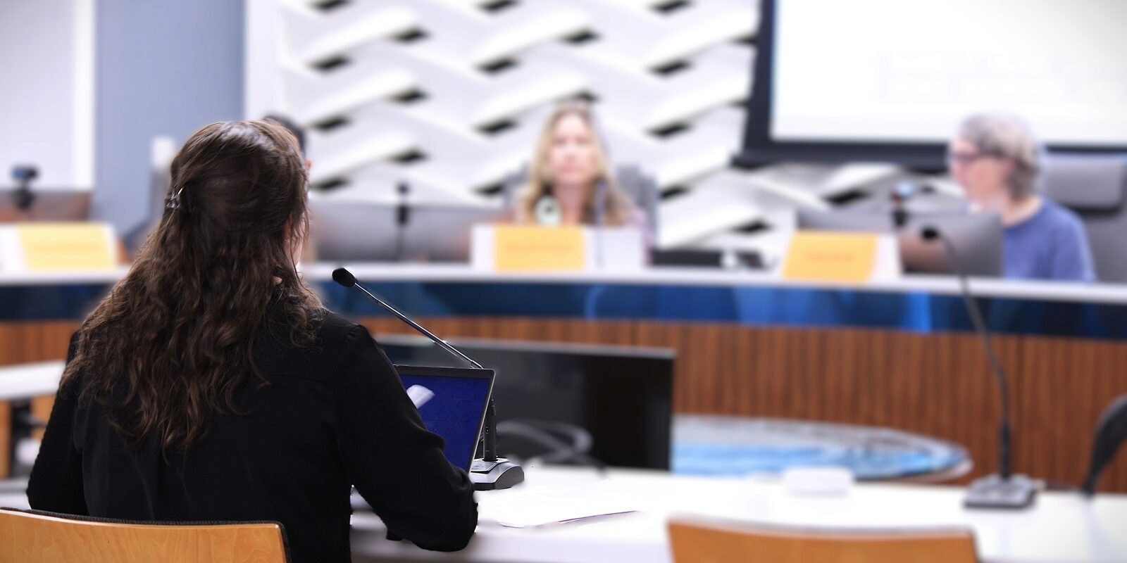 Person seated at a podium with microphone, facing two commissioners at a curved dais.