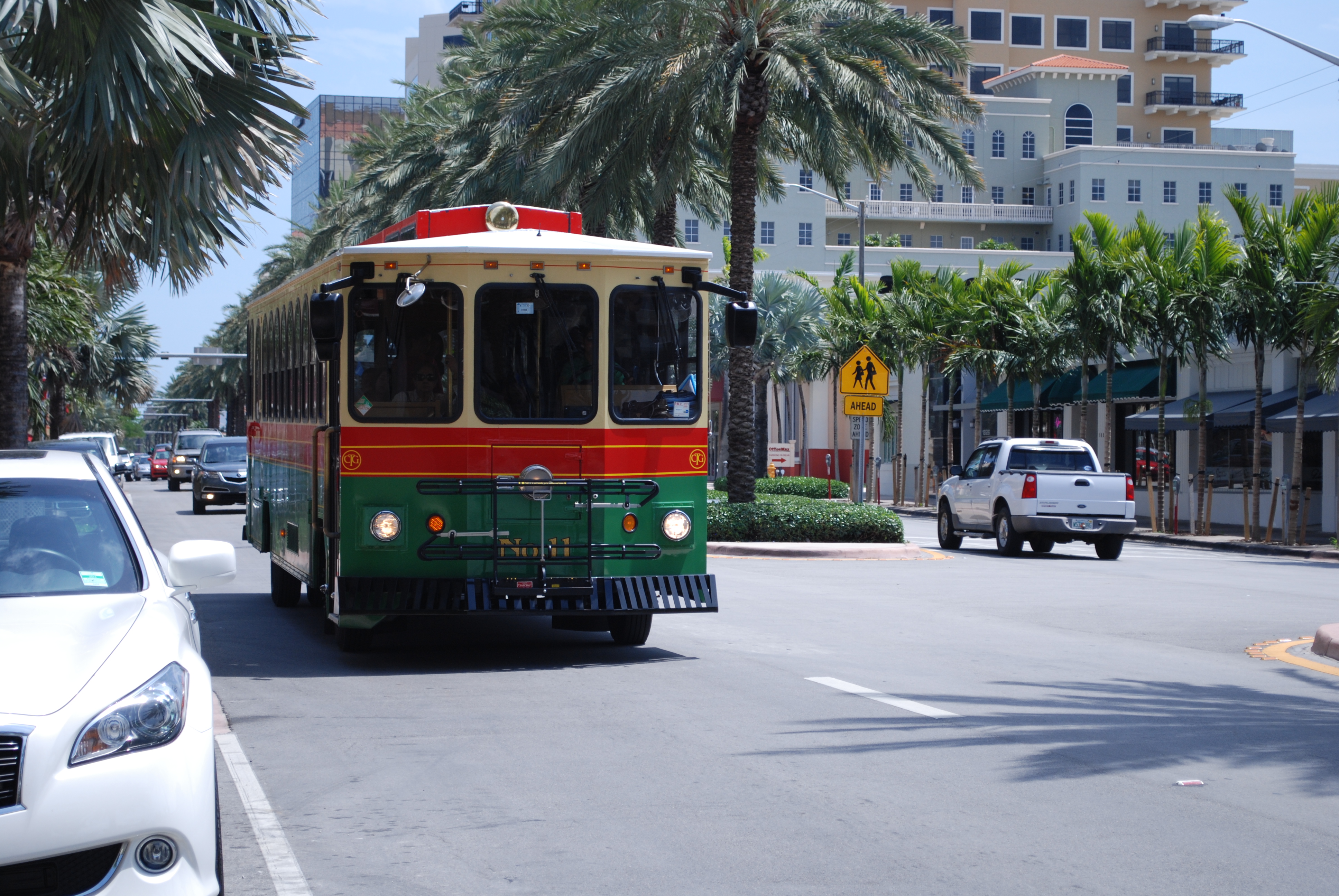 Green, red, and yellow trolley bus traveling along a palm-lined city boulevard.