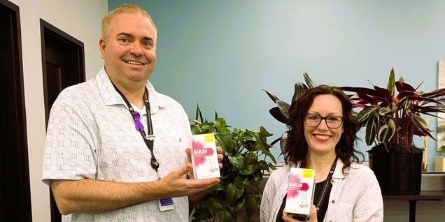 Two smiling people in lanyards hold boxes of naloxone indoors.