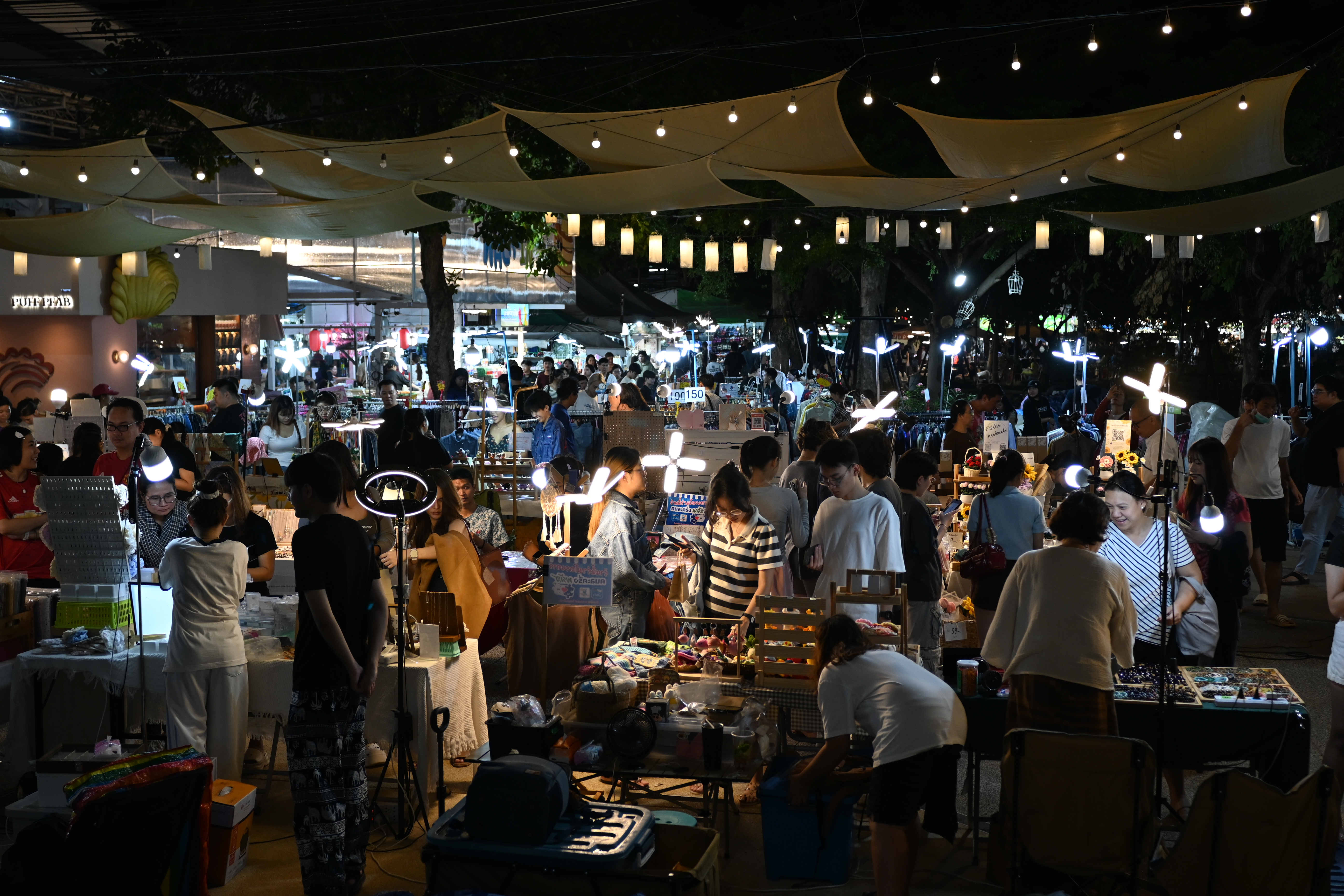 Shoppers browse illuminated vendor stalls under string lights and fabric canopies at a night market.