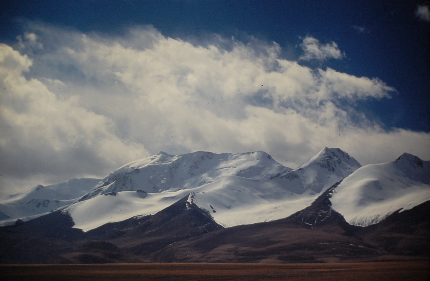 Snow-covered mountain peaks tower above a flat high-altitude plateau under a cloudy sky.