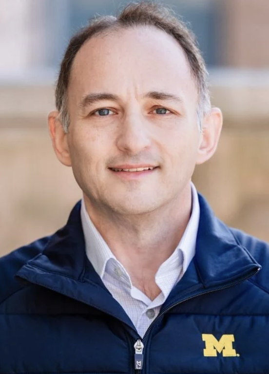 Michael Schostak wears a navy jacket with a maize University of Michigan “M” logo over a light-colored shirt.
