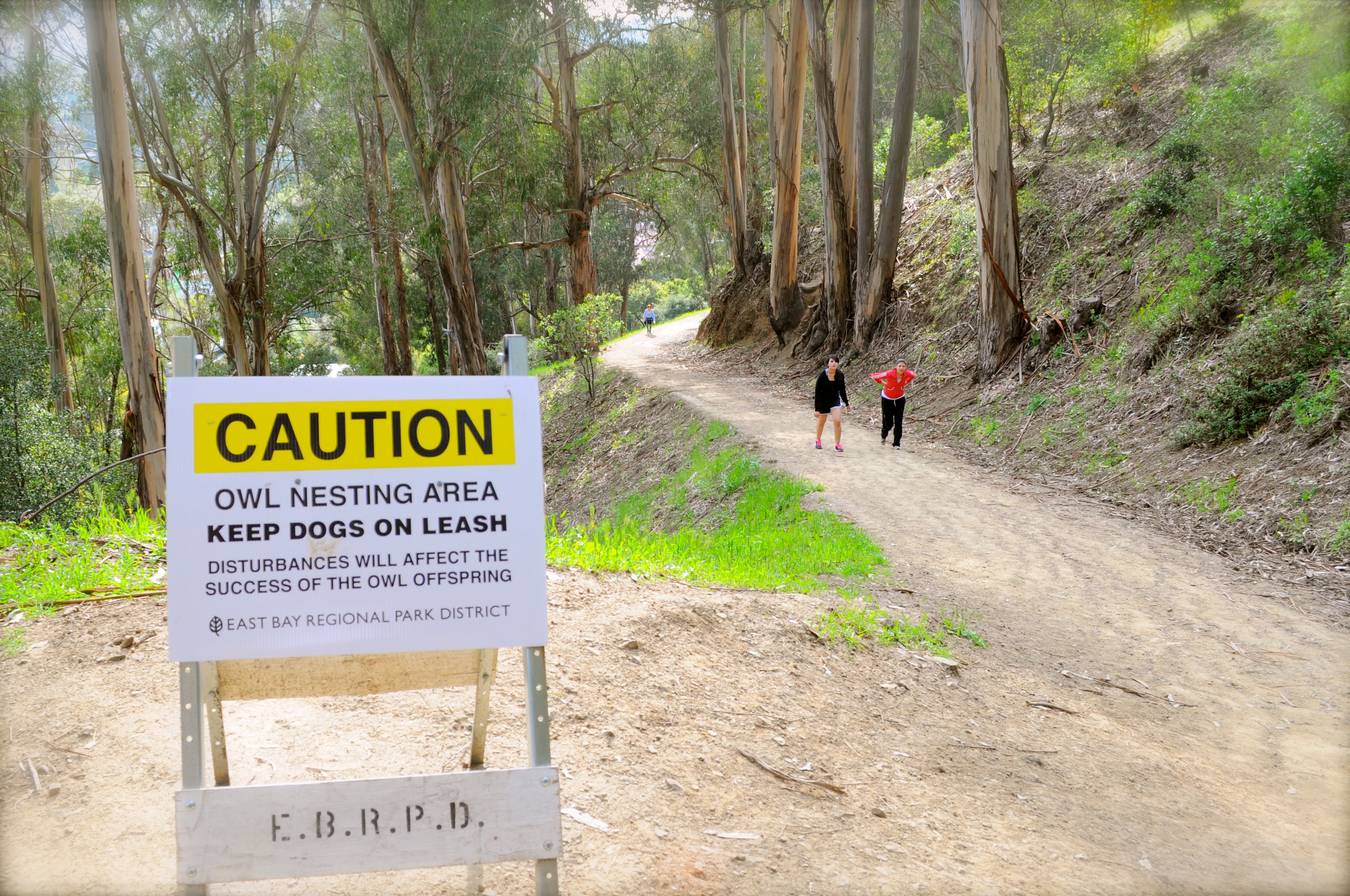 Yellow East Bay Regional Park District sign posted on a dirt trail with hikers in background.