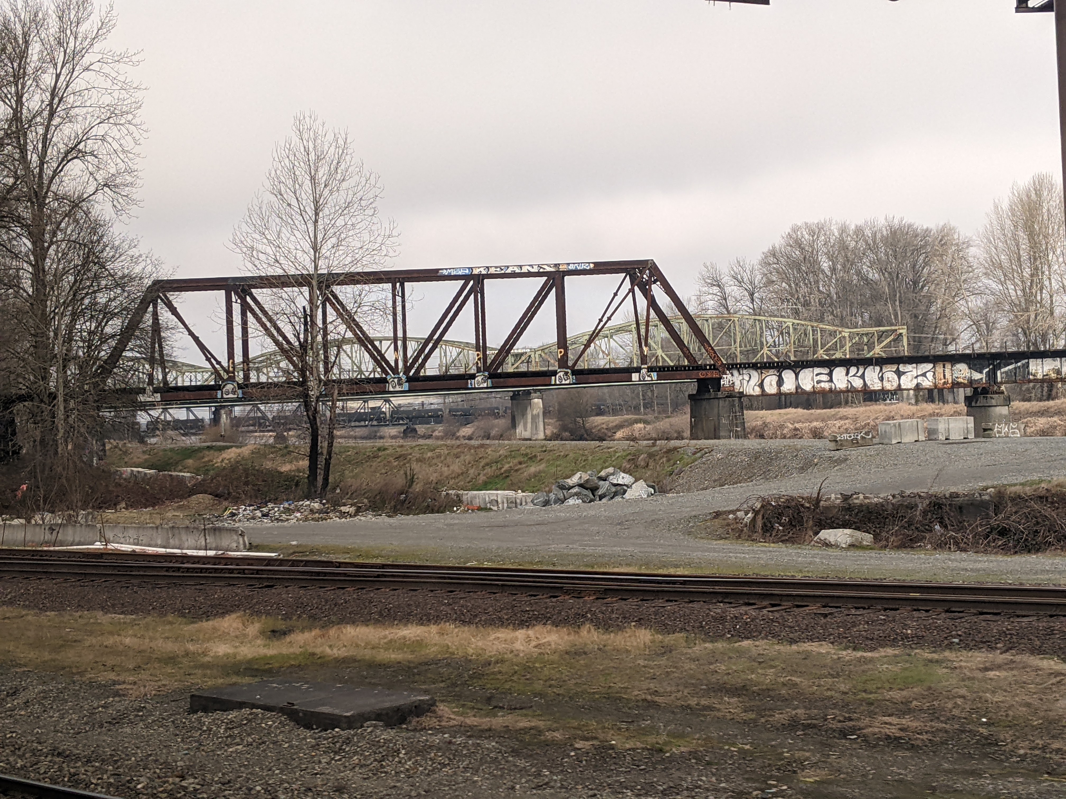 A steel truss bridge spanning a river, viewed from railroad tracks under an overcast sky.