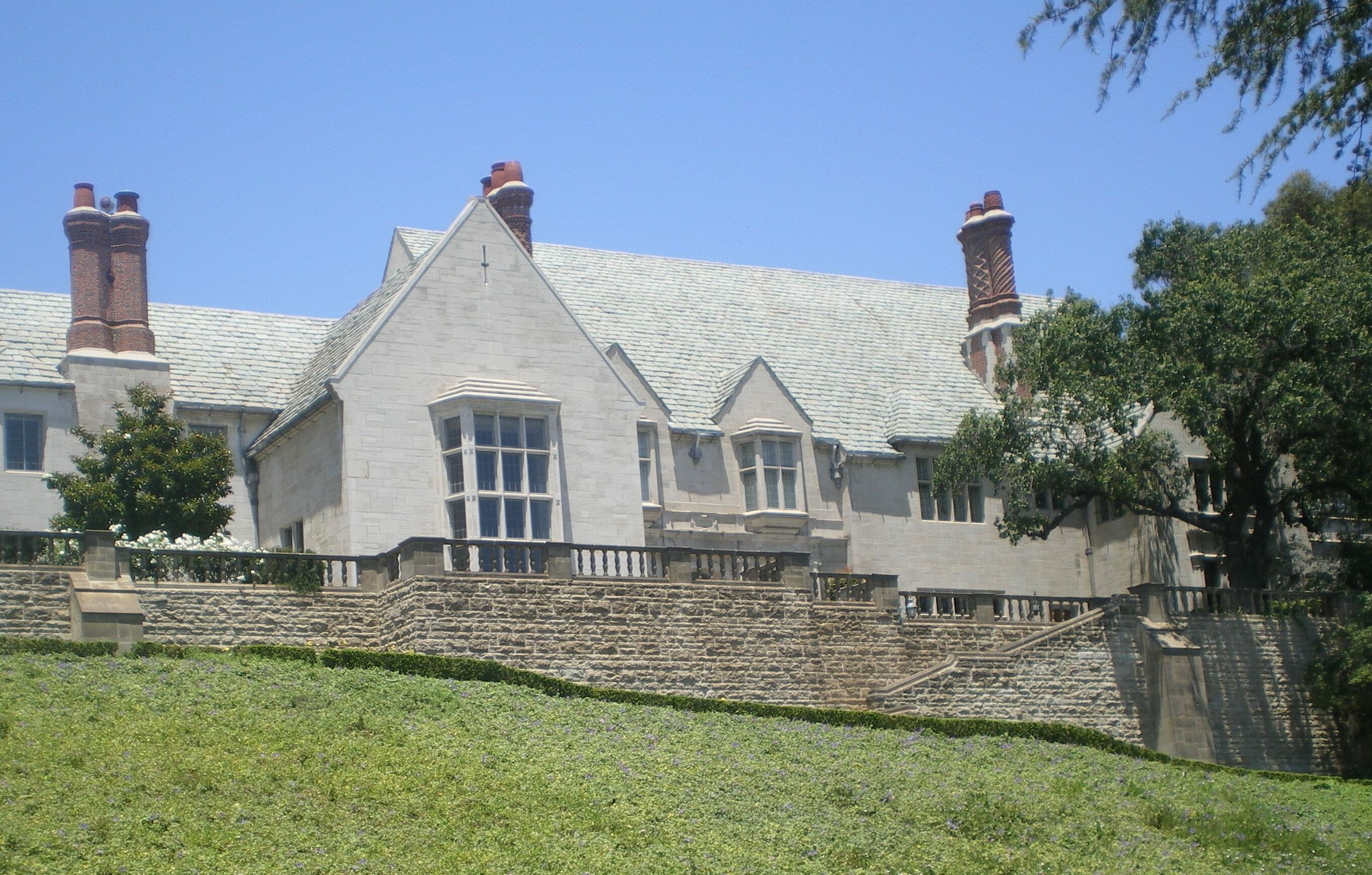 Tudor Revival stone mansion with slate roof and terraced stone walls on a sunny day.