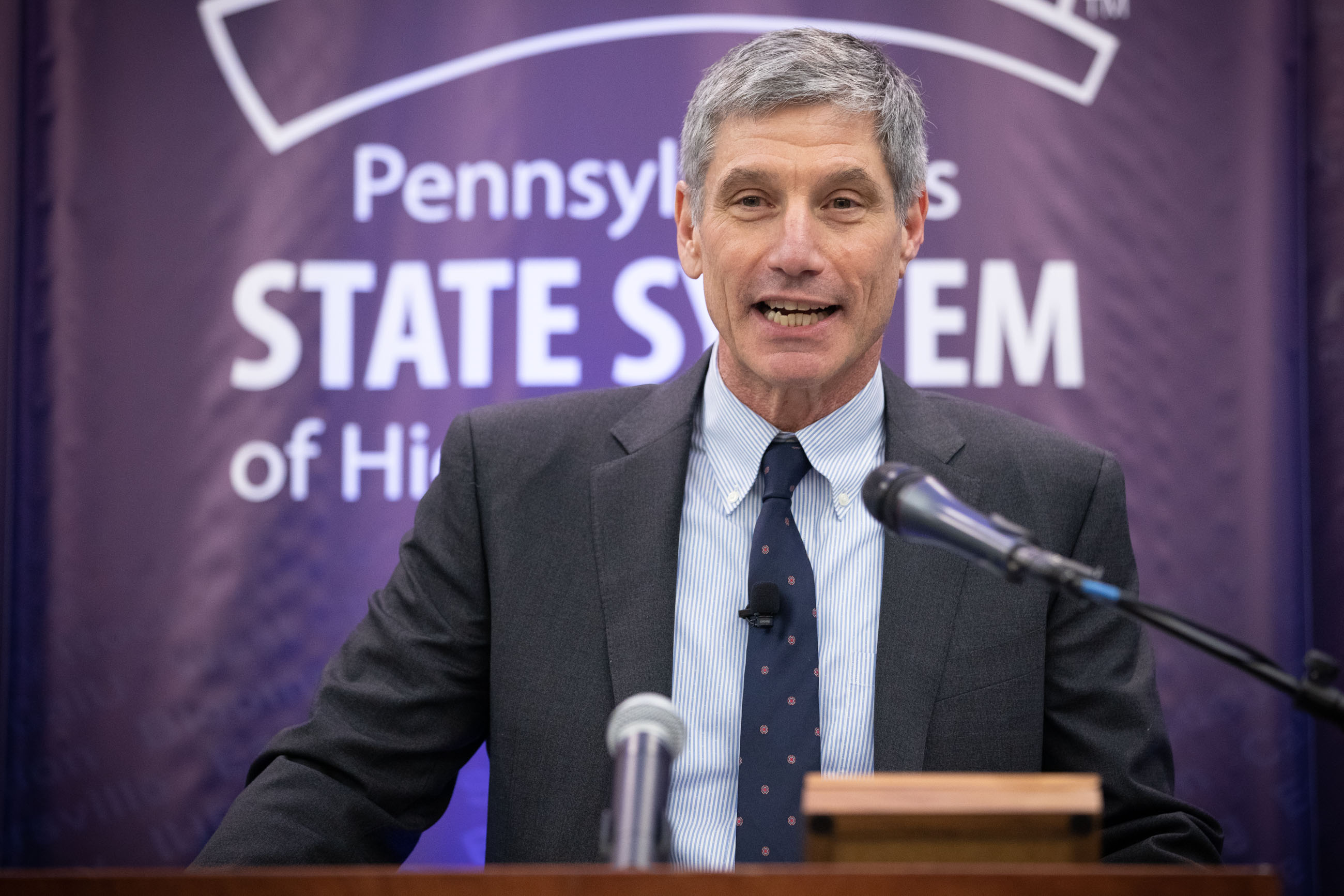 A man in a suit speaks at a podium in front of a higher education system banner.