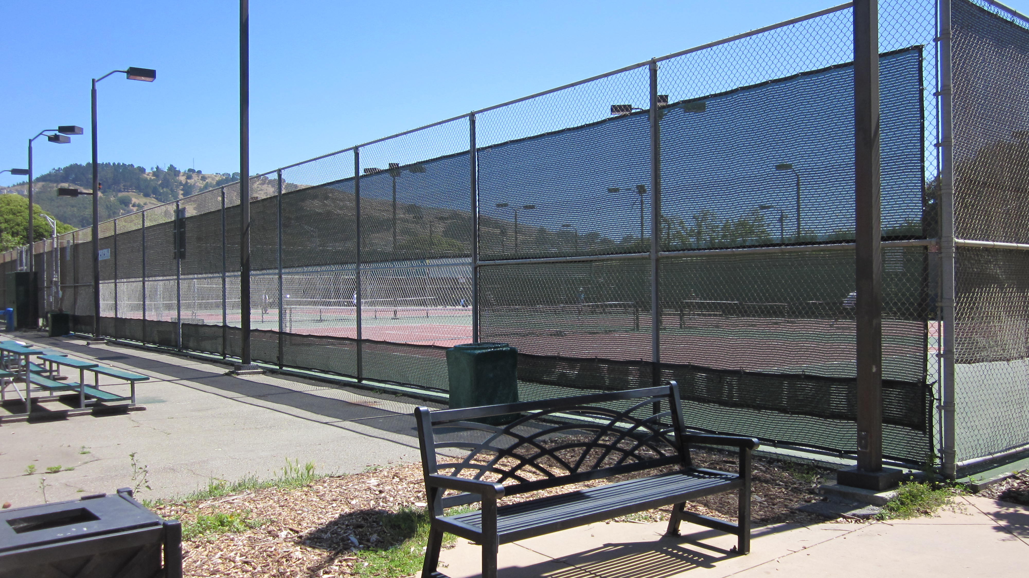 Outdoor tennis courts behind tall chain-link fencing with dark windscreen and light poles.