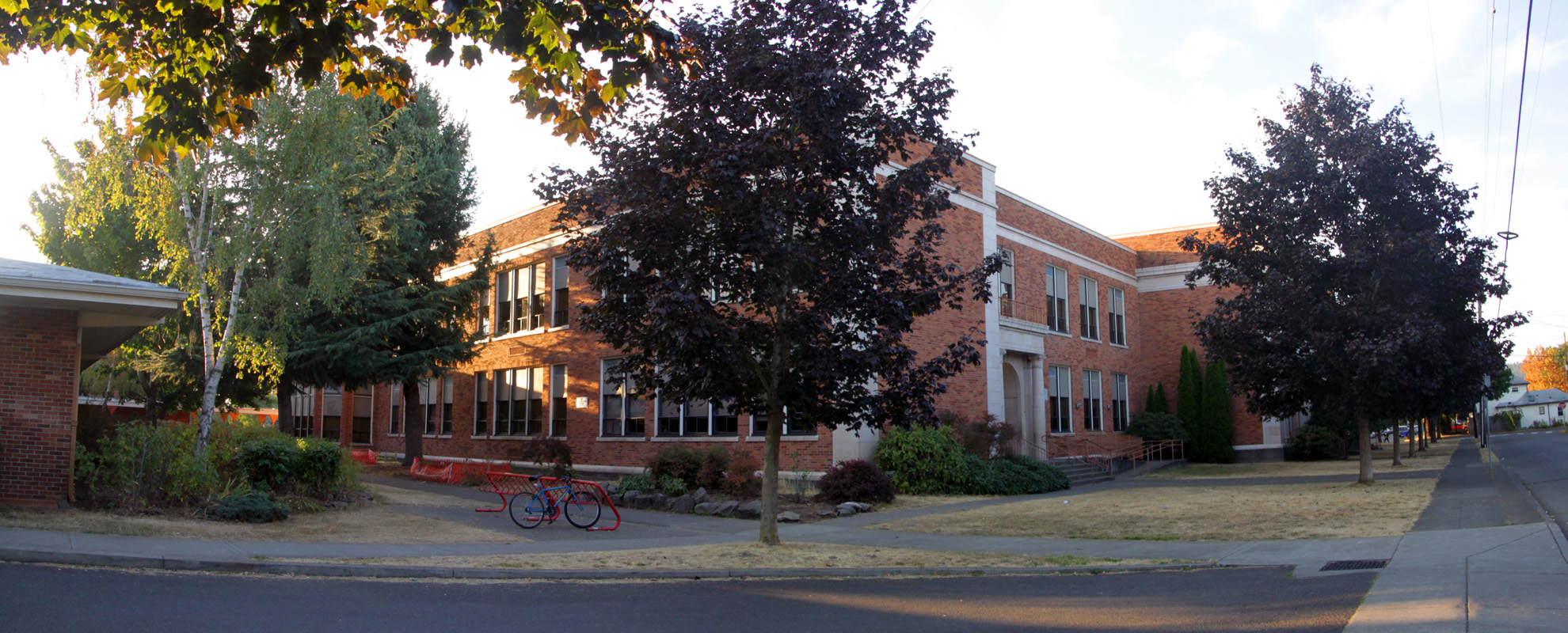 Two-story red brick school building at a street corner, trees and a bicycle out front.
