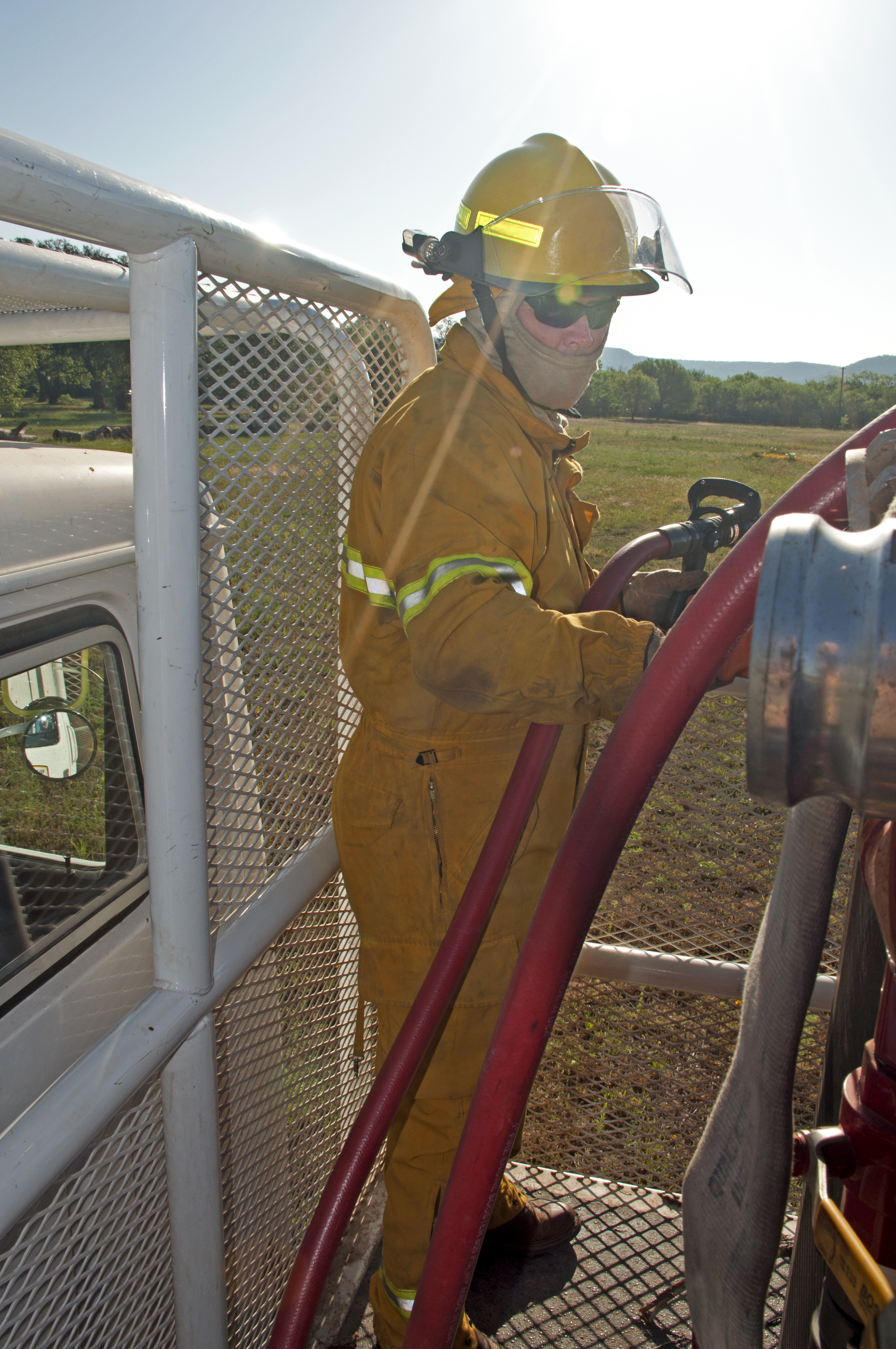 Firefighter in yellow turnout gear and helmet handles a hose line on a red brush truck.