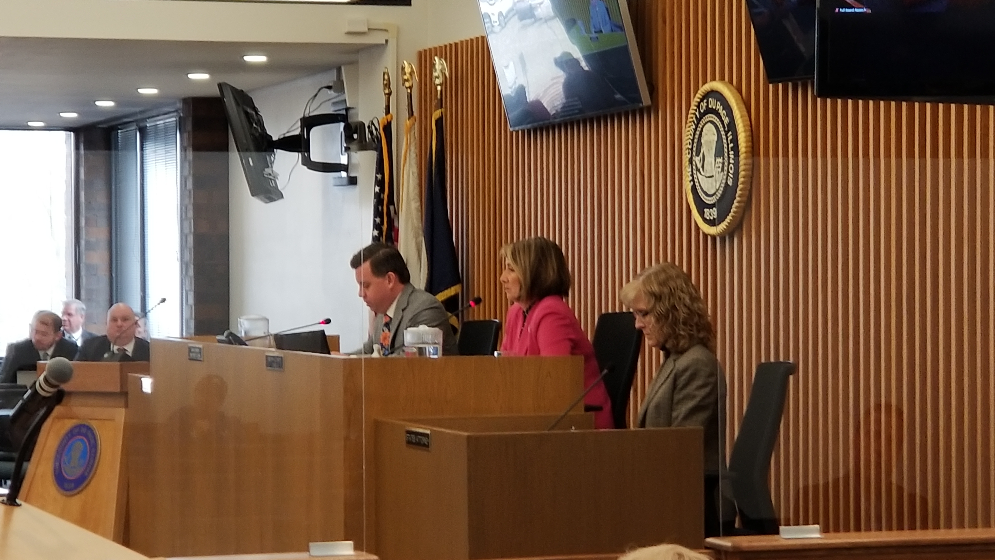Officials seated at a raised dais in a government meeting chamber with flags and a wall seal.