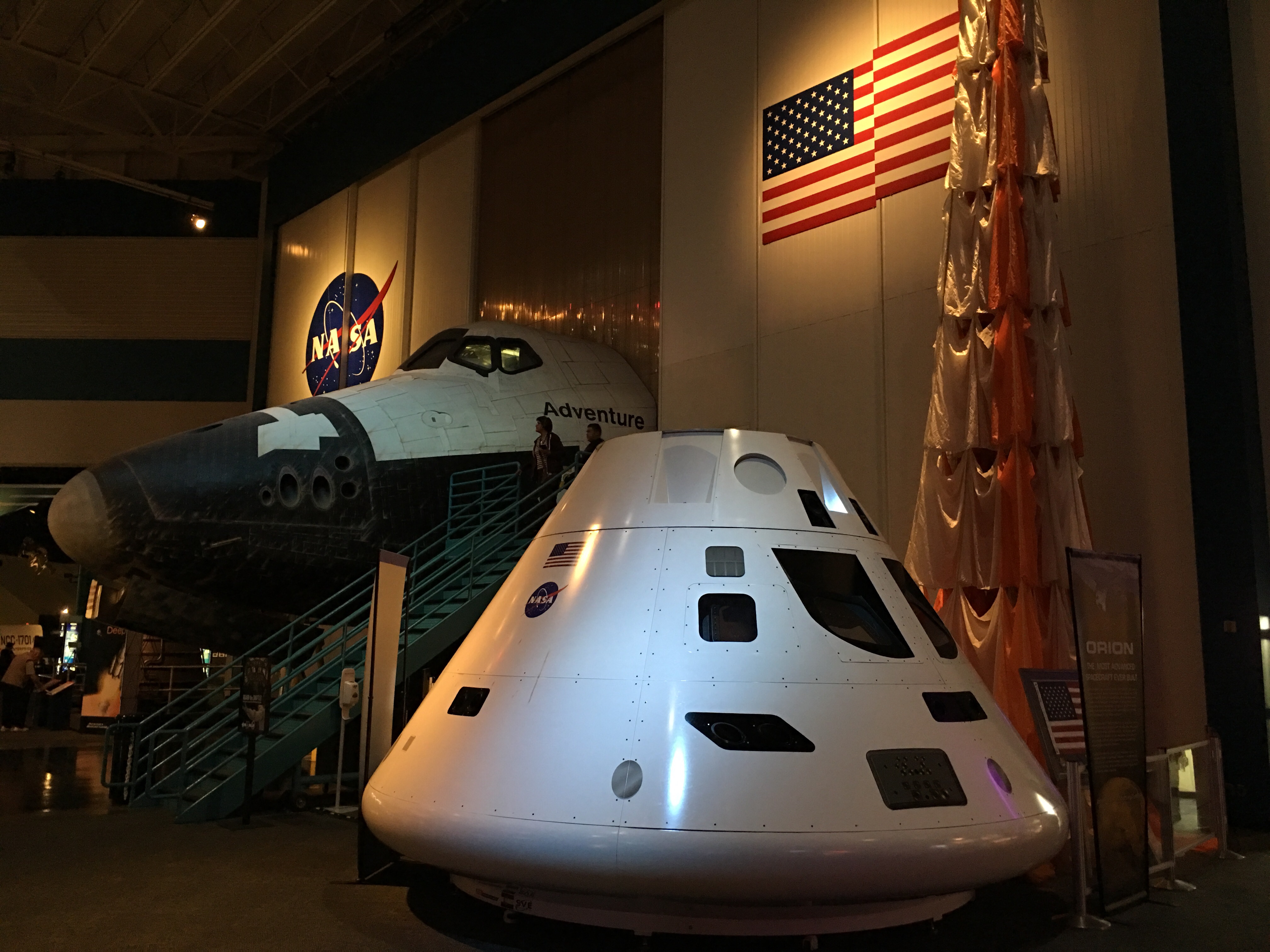 Indoor exhibit hall showing a Space Shuttle orbiter, white crew capsule, and NASA logo.