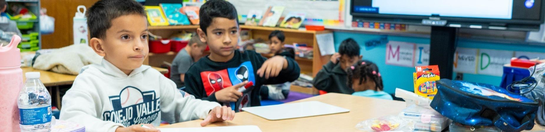Two young students seated at a desk in an elementary school classroom.