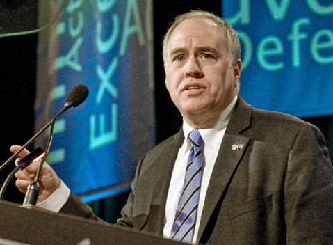 Man in dark suit with striped tie and flag lapel pin speaking at a podium with microphones.
