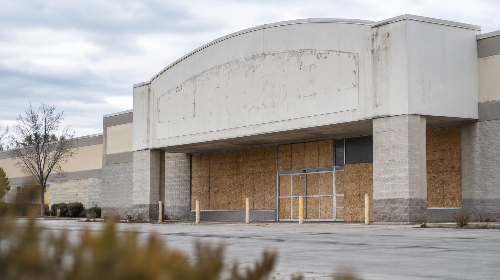 Large vacant retail building with boarded windows and an empty parking lot.