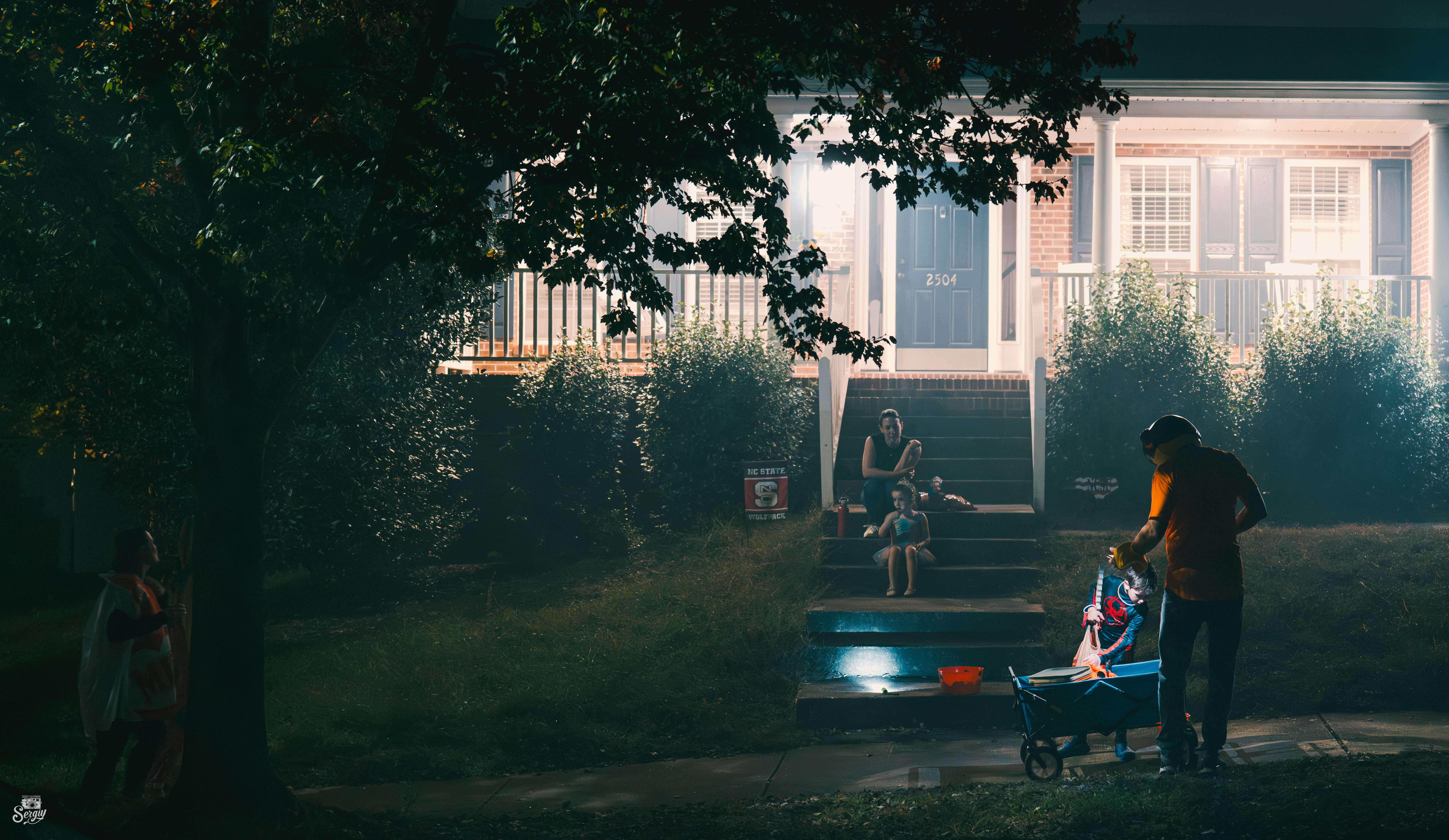 A family with a stroller and children approaches a lit porch on a Halloween evening.