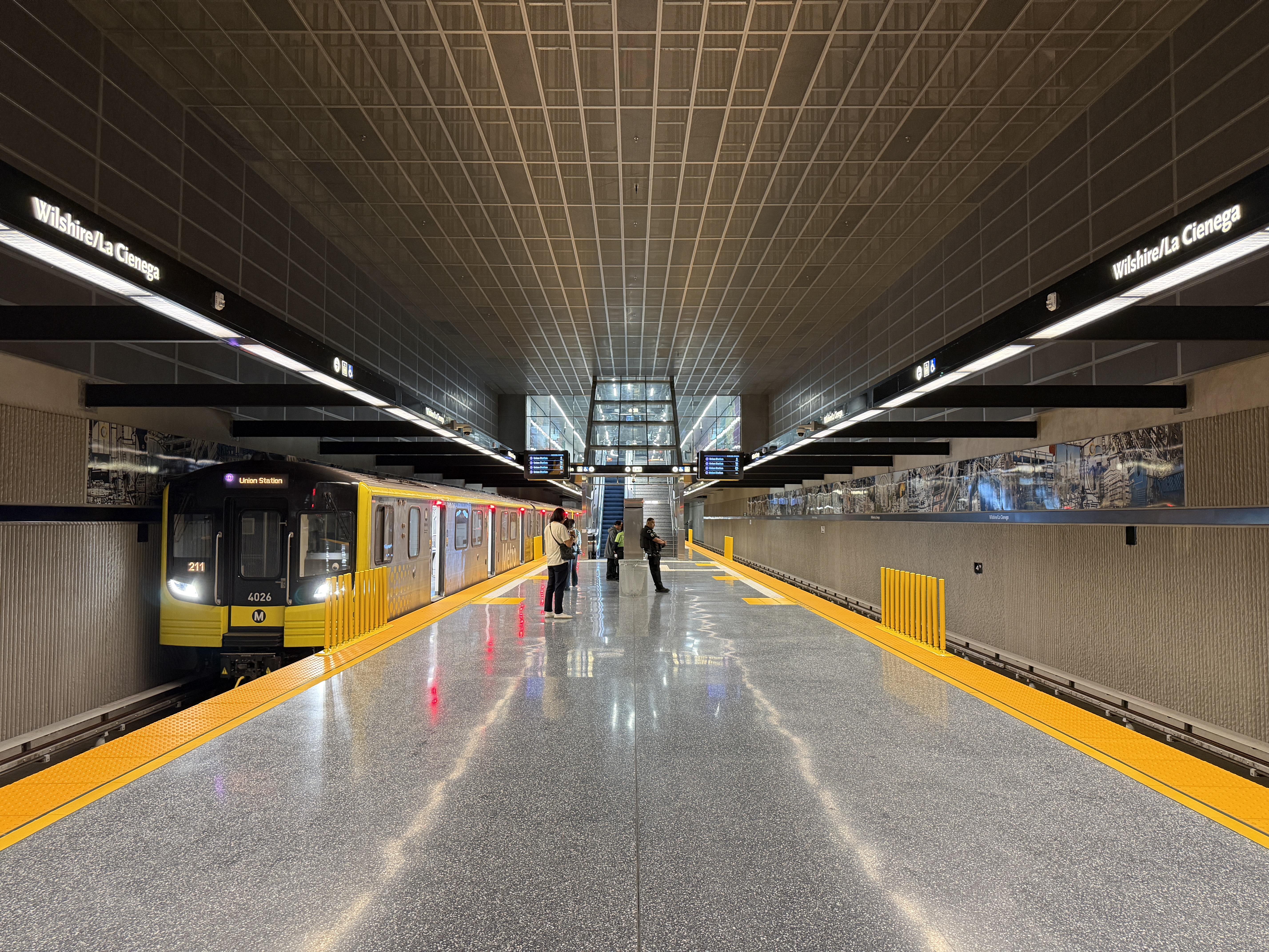 Underground subway platform with a yellow Metro train and passengers under a grid ceiling.