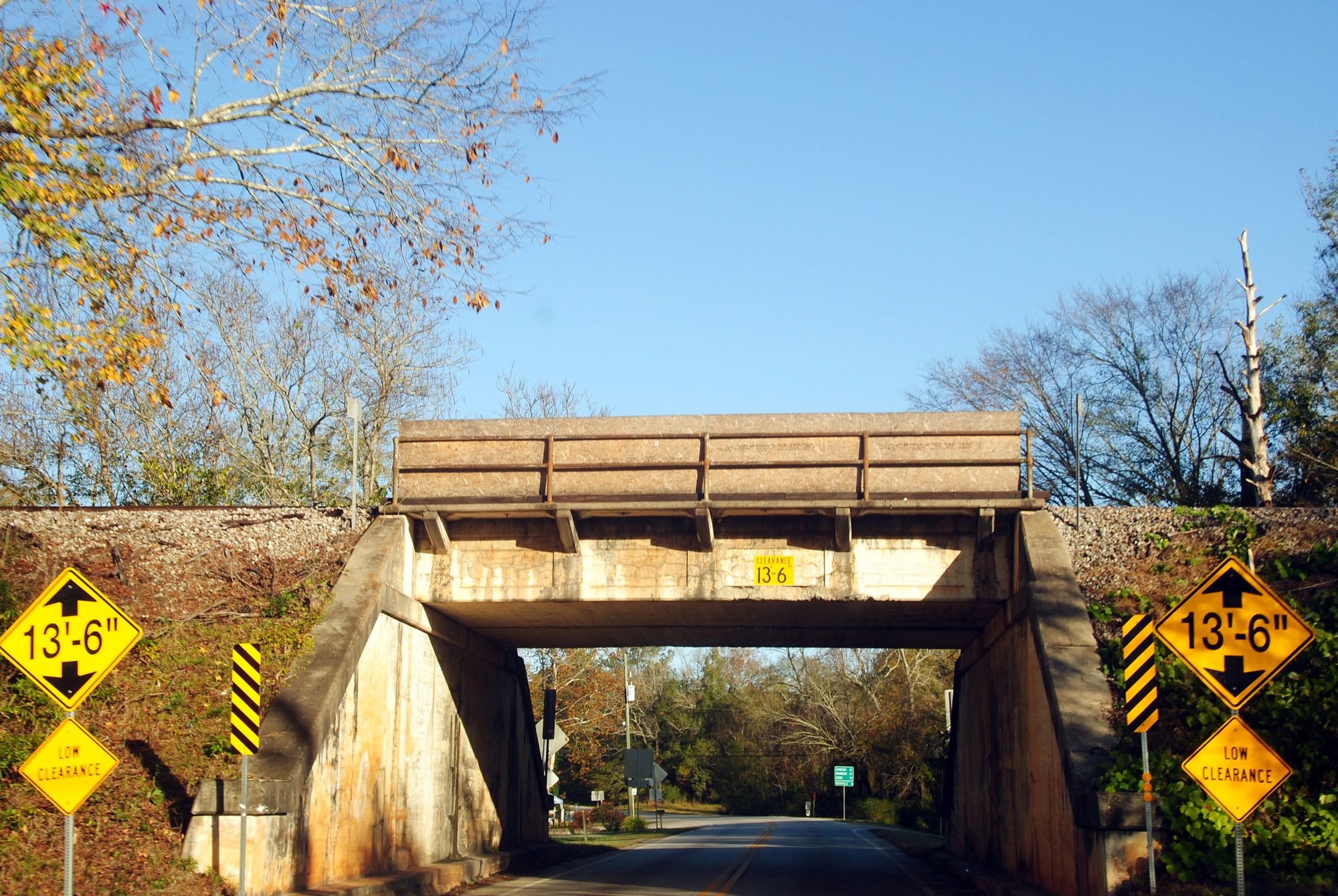 Railway bridge over country road with low clearance. Autumn foliage surrounds the scene on a bright day.