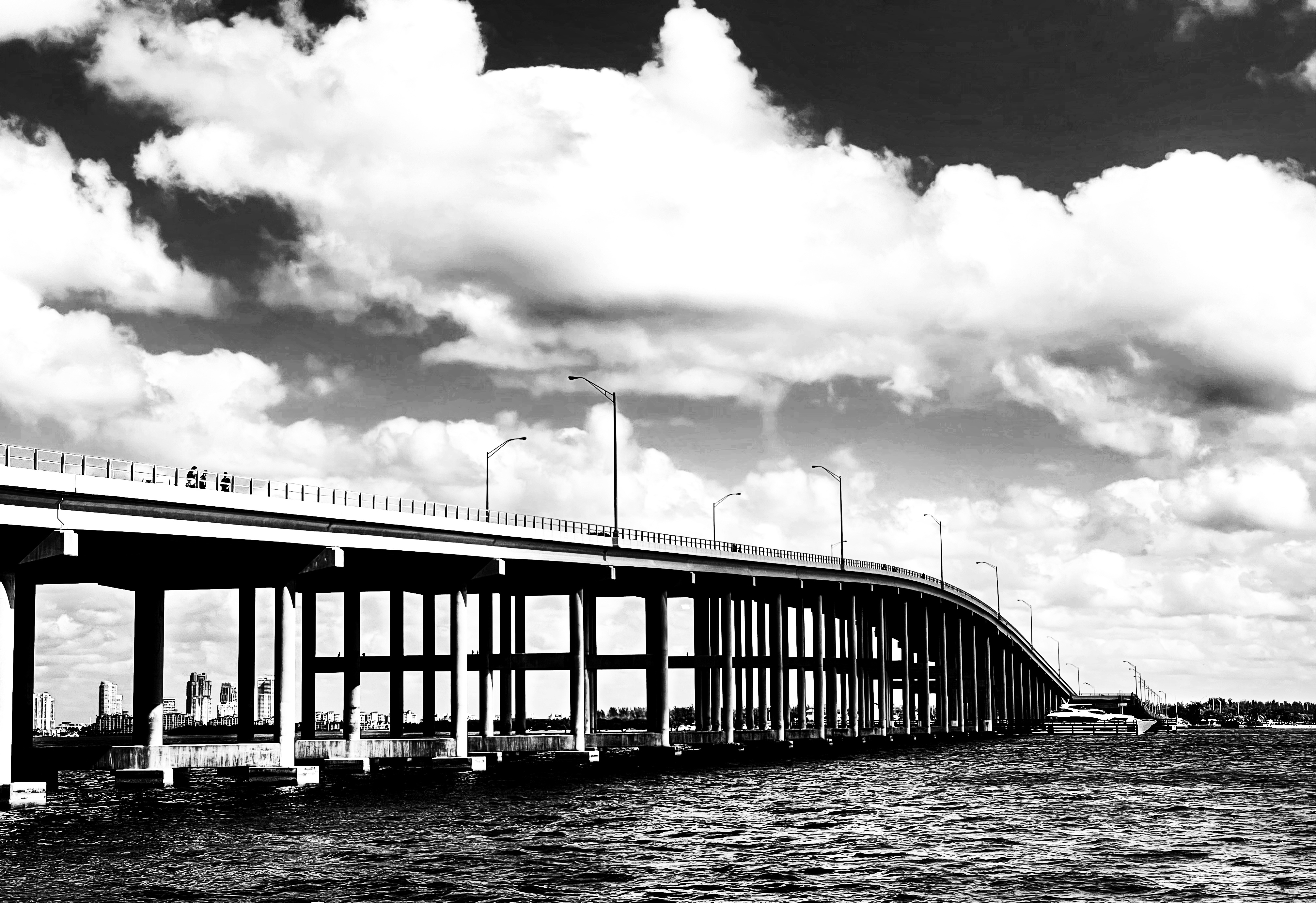 Long elevated bridge spanning open water under a dramatic cloudy sky, in black and white.