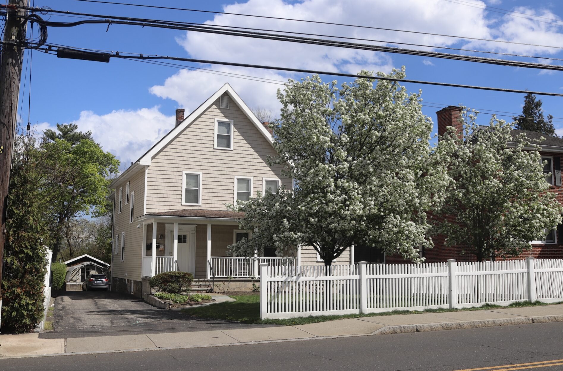 A beige two-story house with a white picket fence and flowering tree in the front yard.