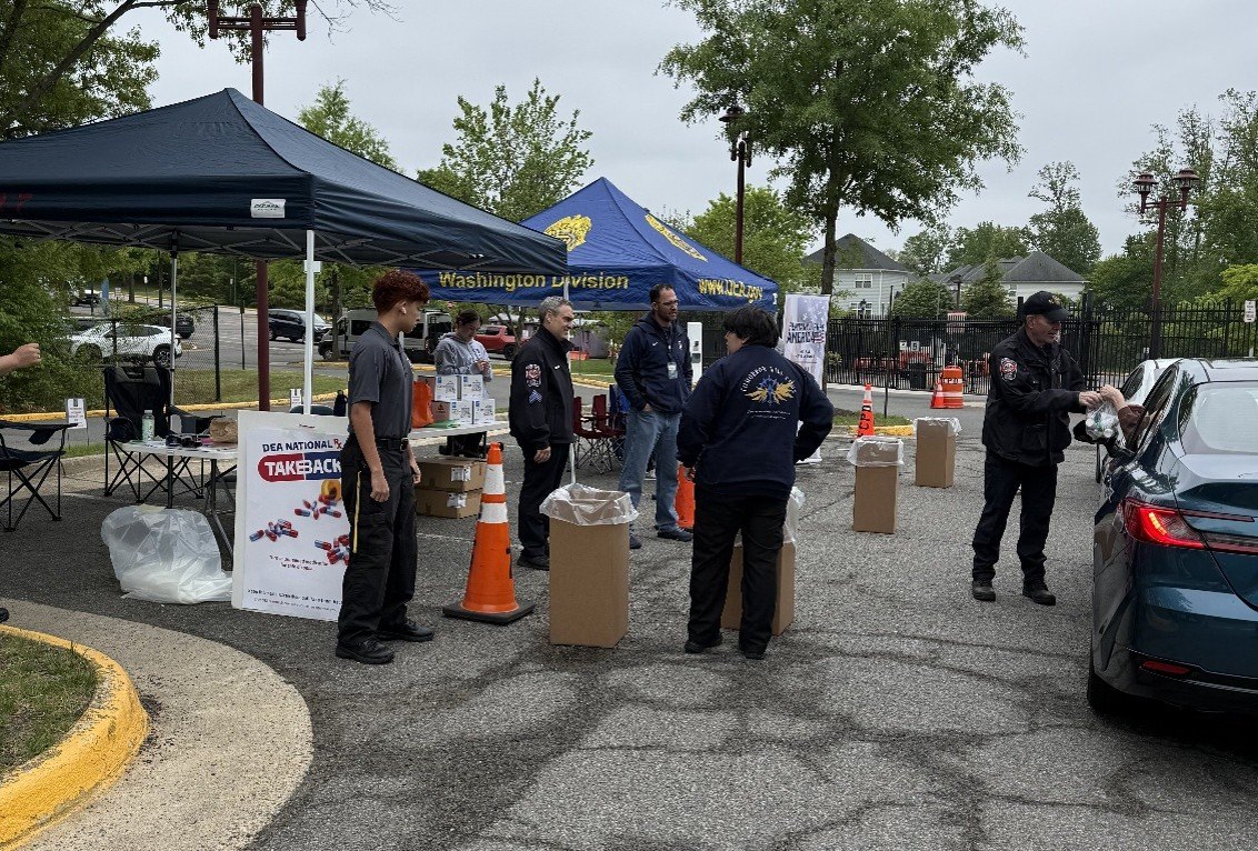 Outdoor drug take-back collection site with a blue DEA tent, orange cones, and cardboard boxes.