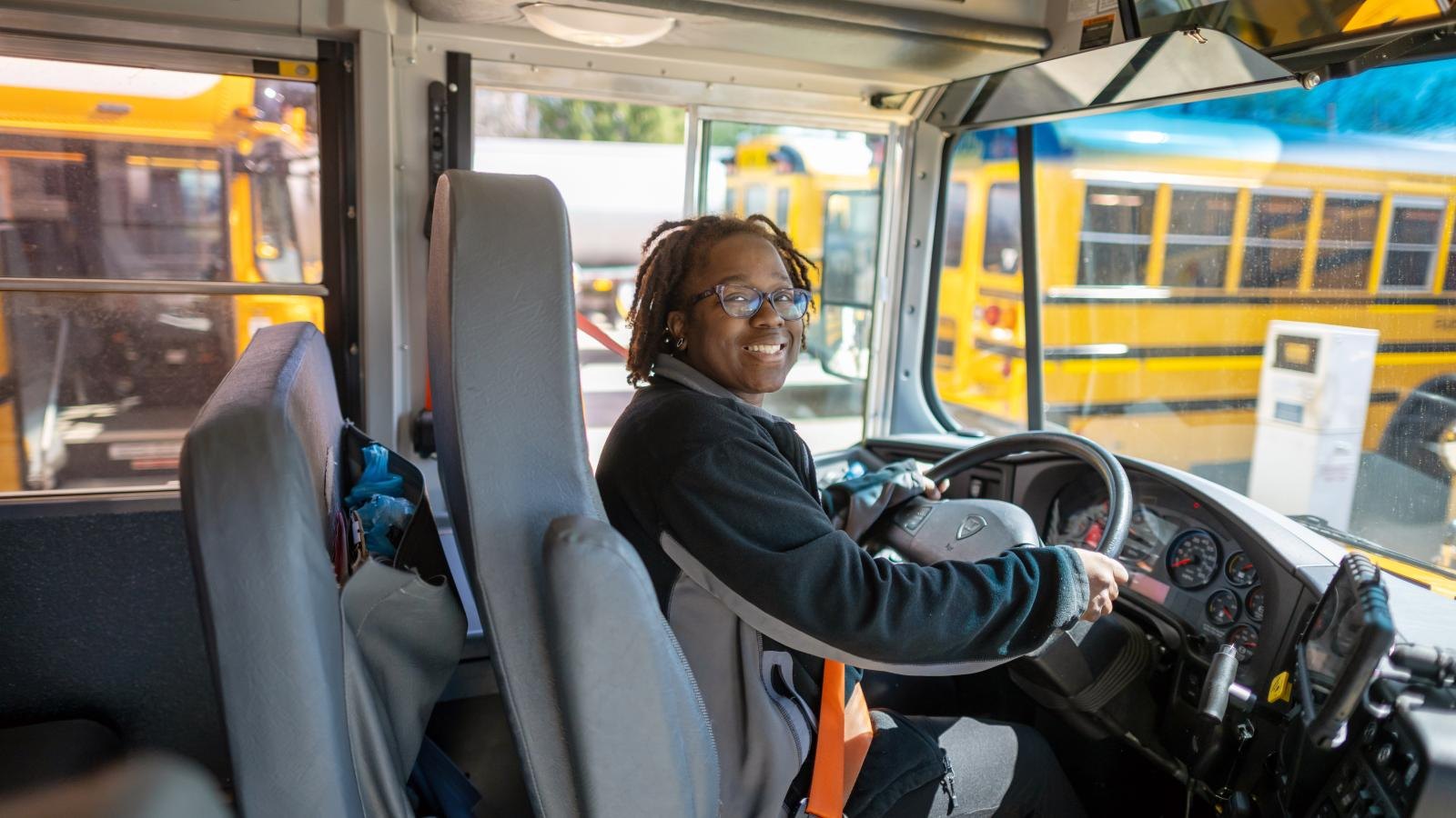 A smiling person with dreadlocks and glasses seated at a school bus steering wheel.