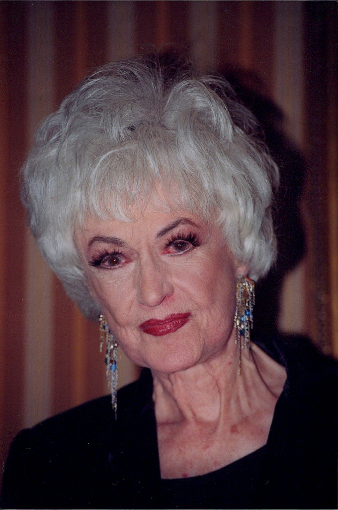 A woman with short white hair, red lipstick, and dangling earrings in a black top.