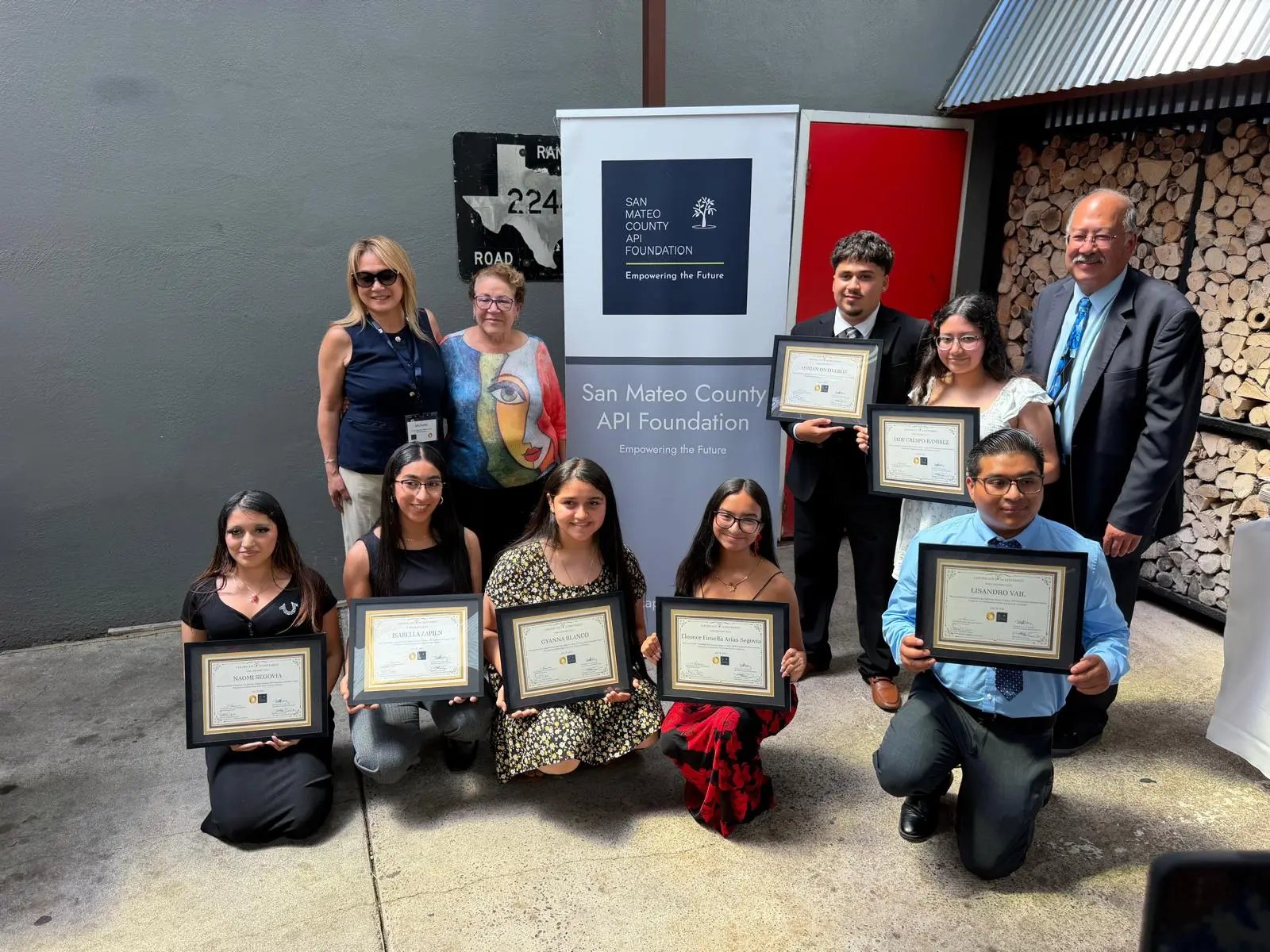 Group of students and adults holding framed certificates in front of a nonprofit banner.