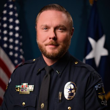 Police officer in dark uniform with badge and service ribbons, posed before American and Texas flags.
