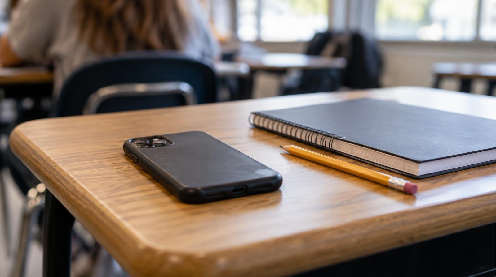 A smartphone face-down on a wooden student desk next to a closed notebook and pencil.