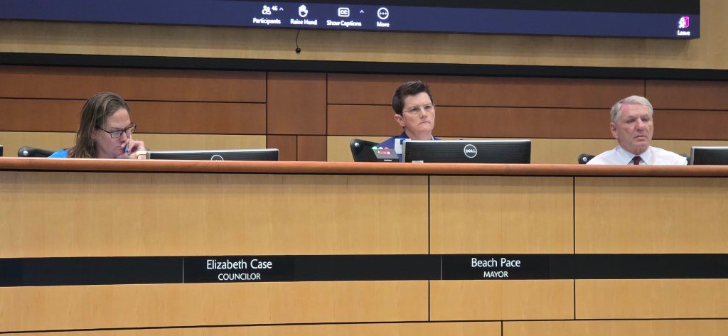 Three council members seated at a raised dais with monitors and name plates during a city council meeting.