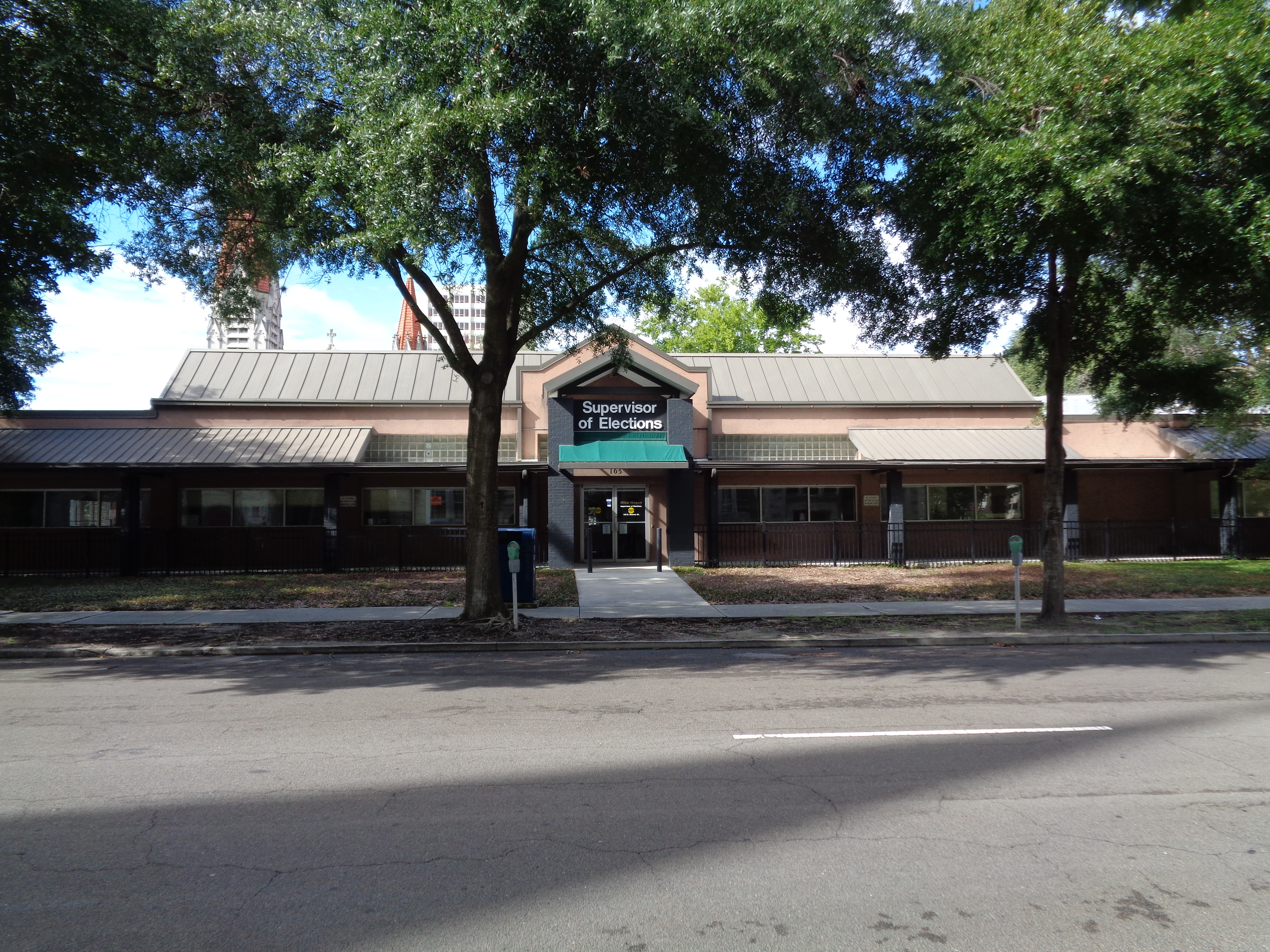 Single-story brown building with a teal sign reading Supervisor of Elections, surrounded by trees.