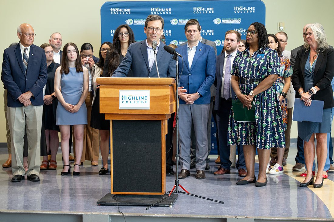 Man in dark suit speaking at a Highline College podium with supporters standing behind him.