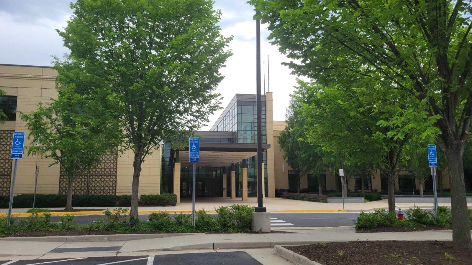 Modern school building with glass entrance and beige exterior surrounded by trees and an empty parking lot.