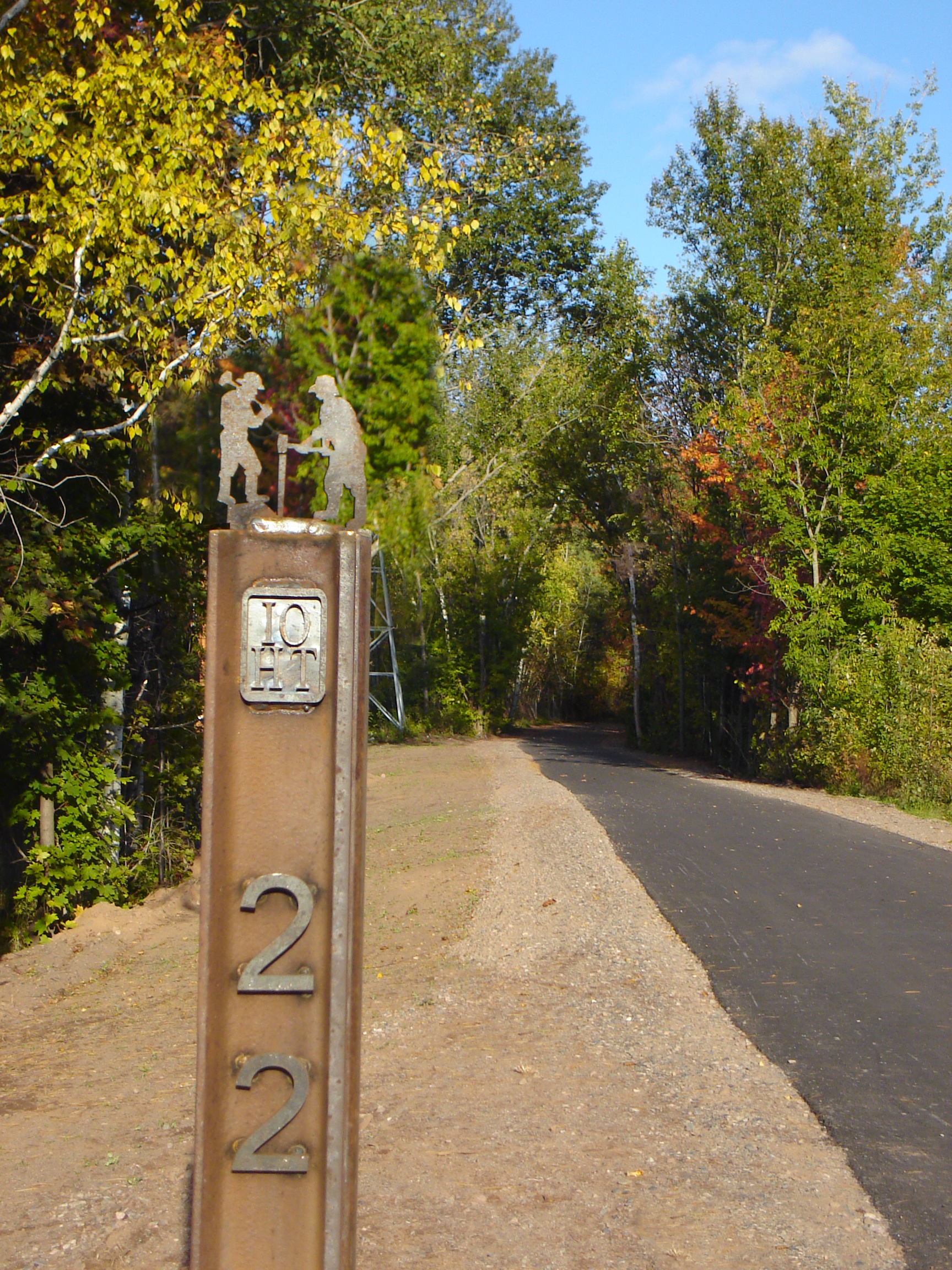 Mile marker post for the Iron Ore Heritage Trail beside a paved path lined with autumn trees.