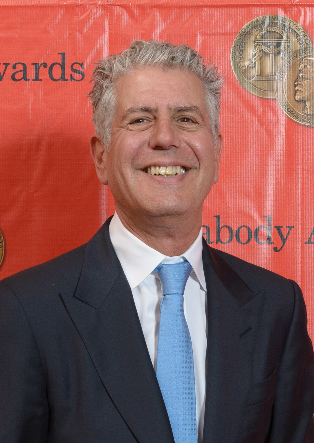 Smiling man with gray hair in a dark suit before a red awards ceremony backdrop.