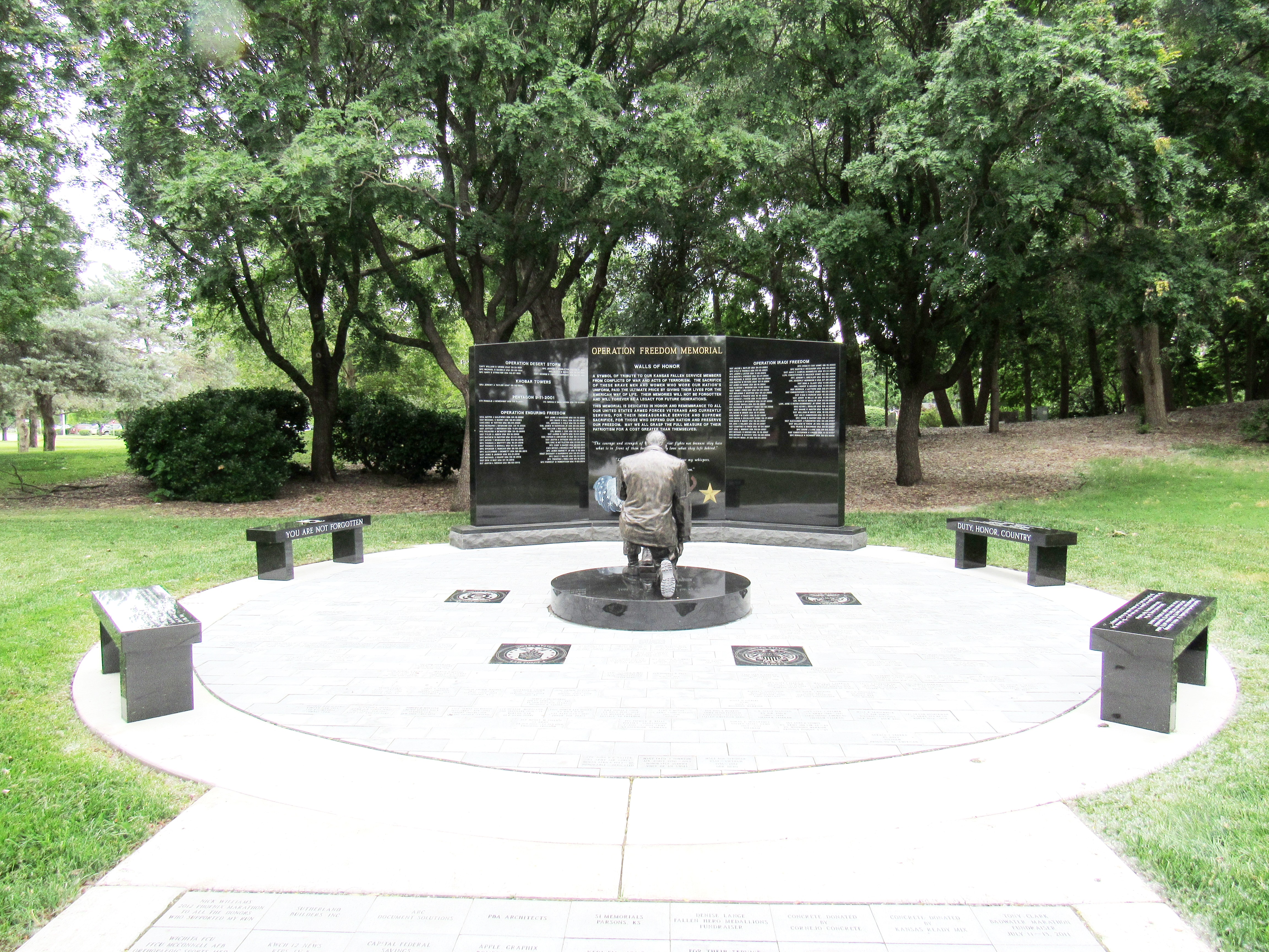 Bronze kneeling soldier statue at the center of a circular memorial plaza with granite panels.