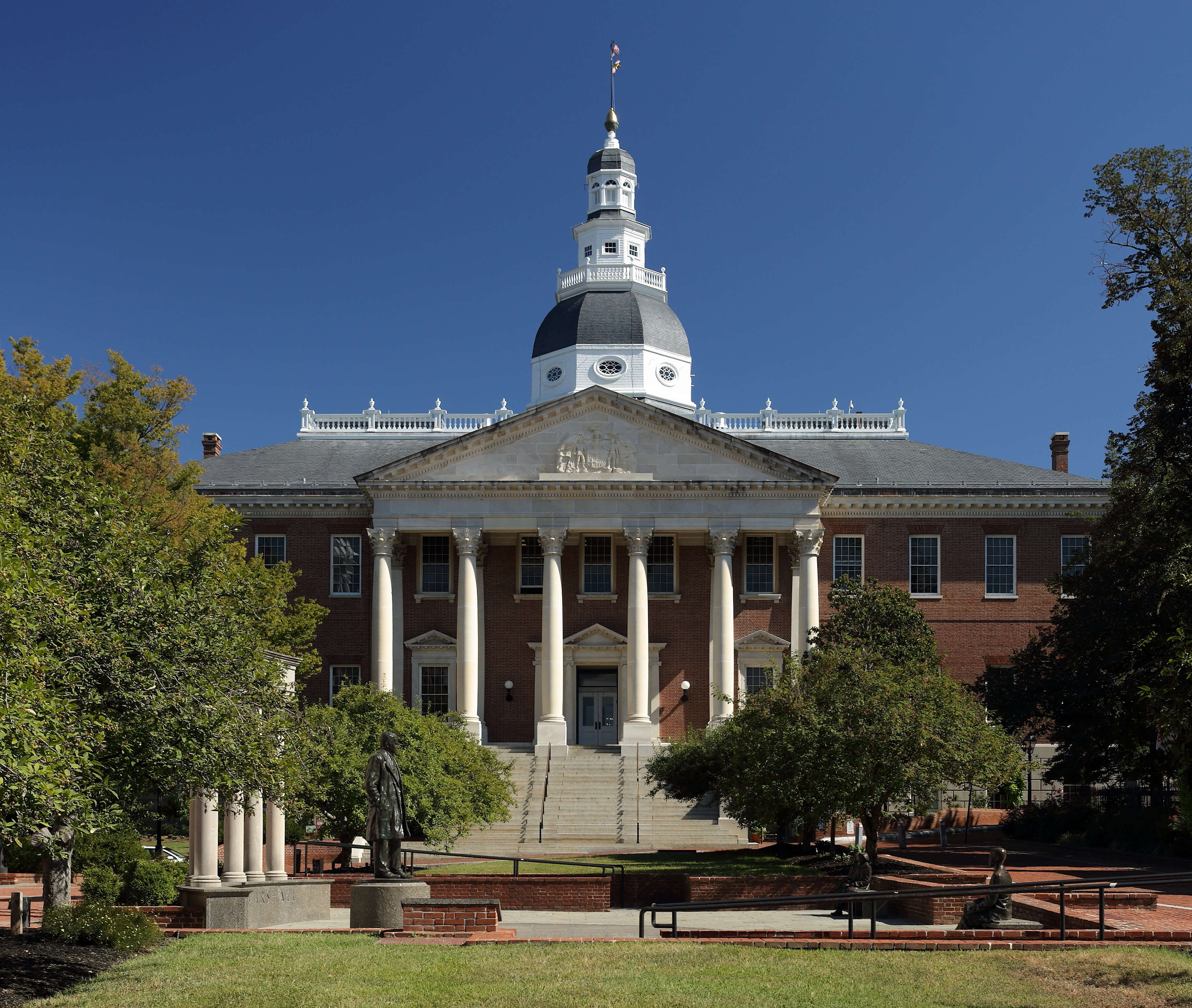 Red brick government building with large white dome and columned entrance, flanked by trees.