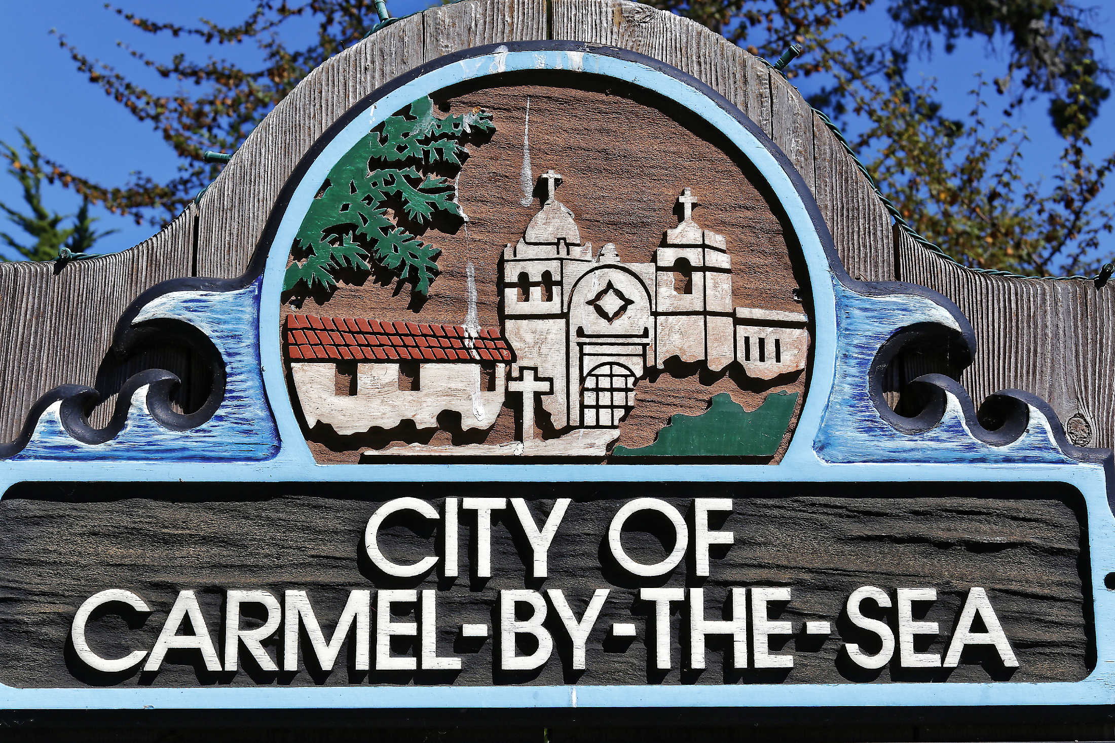 Carved wooden sign reading City of Carmel-by-the-Sea against blue sky and trees.
