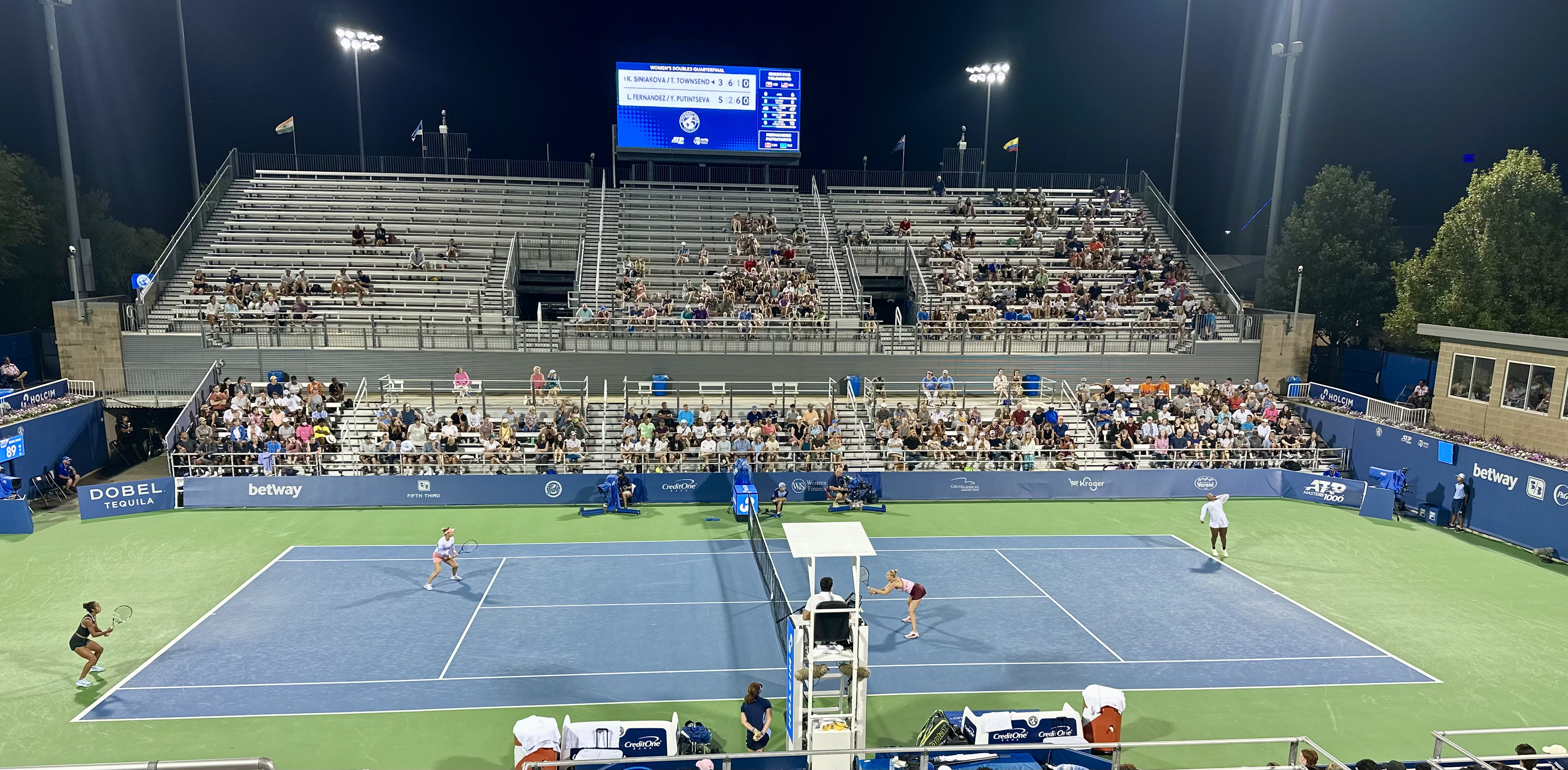 Night tennis match under floodlights on a blue hard court inside a packed stadium.