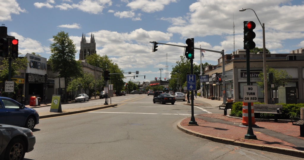 Busy main street intersection with storefronts, traffic signals, and a church steeple in the distance.