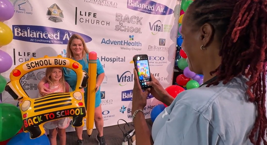 A child and adult pose in a school bus photo prop at a back-to-school event with sponsor banners behind them.