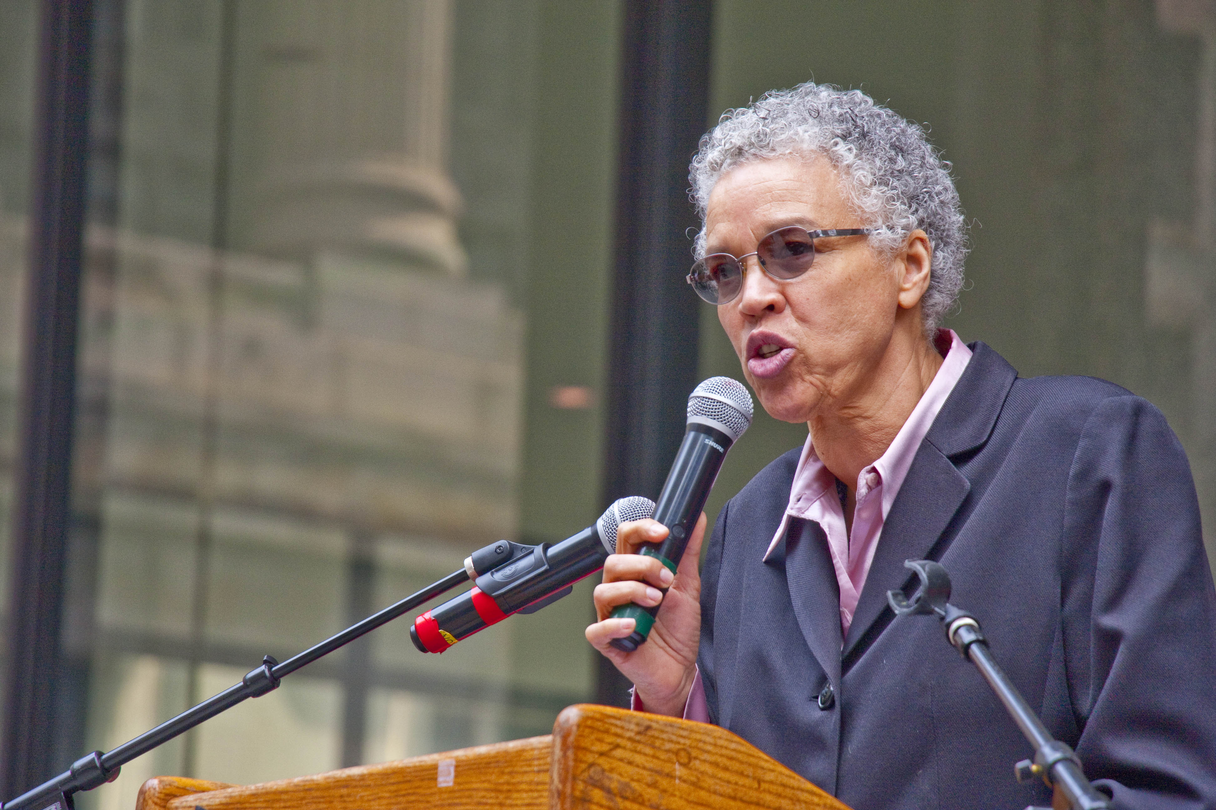 Woman with gray hair and glasses speaking into a microphone at an outdoor podium.