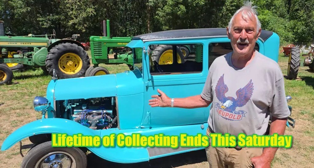 Older man in gray t-shirt standing beside a light blue hot rod with its hood open, green tractor in background.