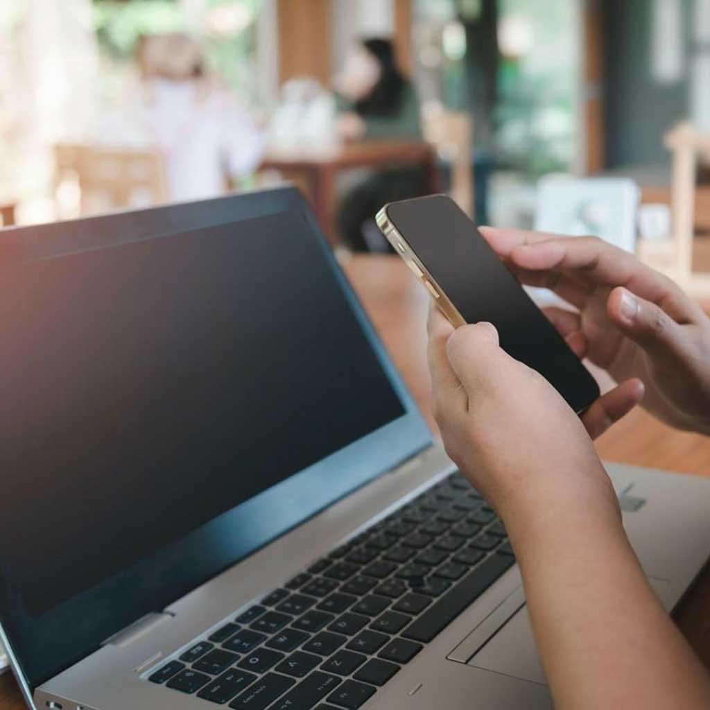 Hands holding a smartphone in front of an open laptop in a public space.