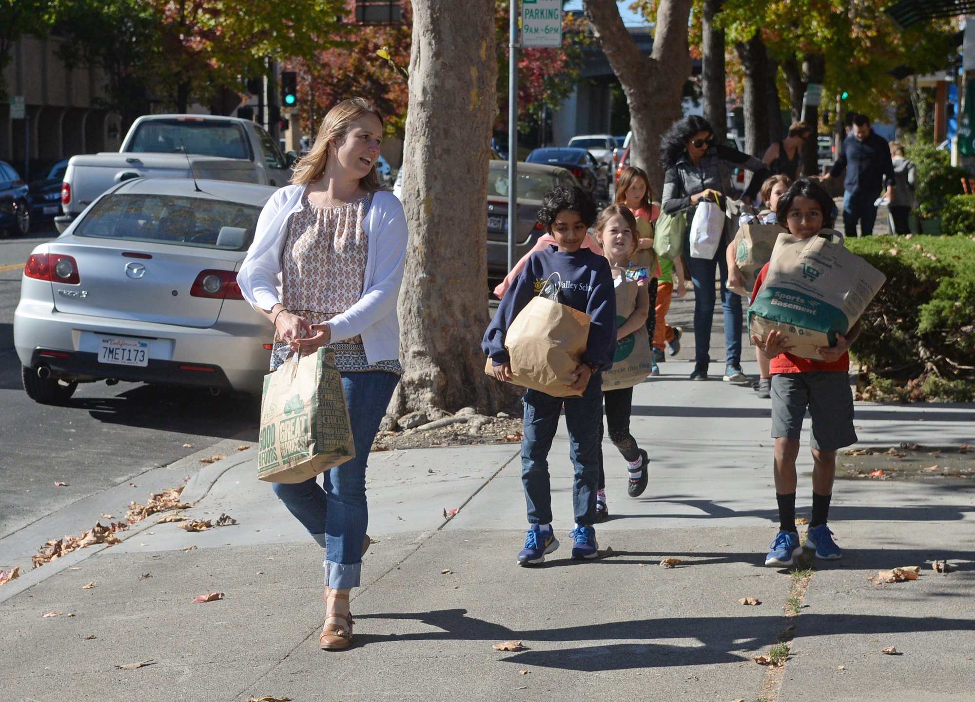 A woman carrying a paper bag walks along a tree-lined sidewalk with several children also carrying bags.